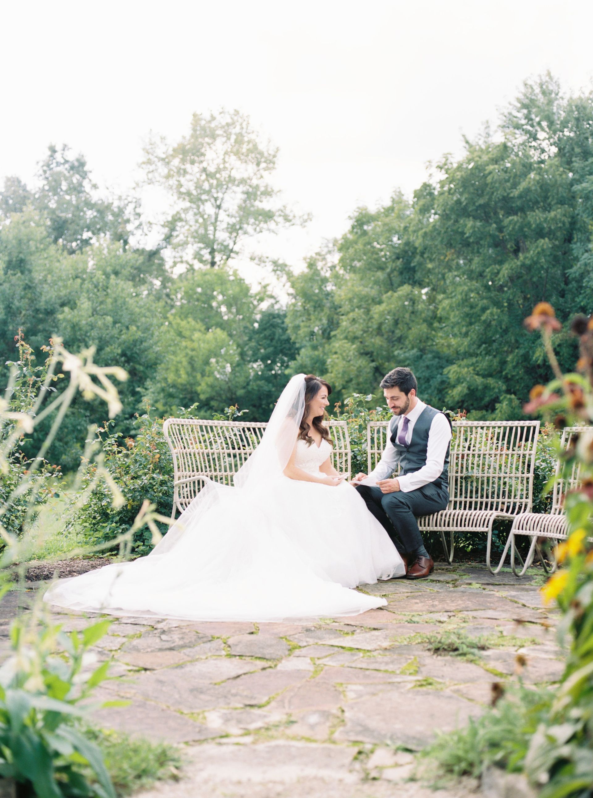 Bride and groom on a bench in a garden; she's in a white gown and veil, he wears a vest.