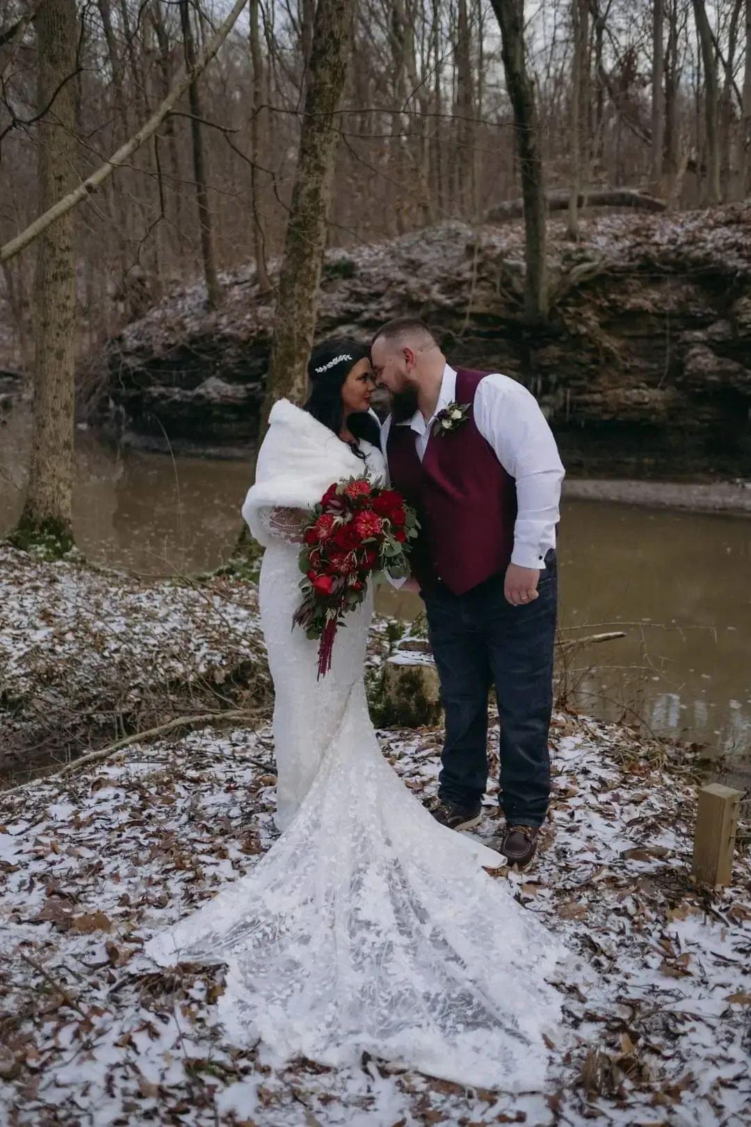 Couple in wedding attire embrace near a creek, surrounded by trees and snow. She holds flowers. He wears a maroon vest.