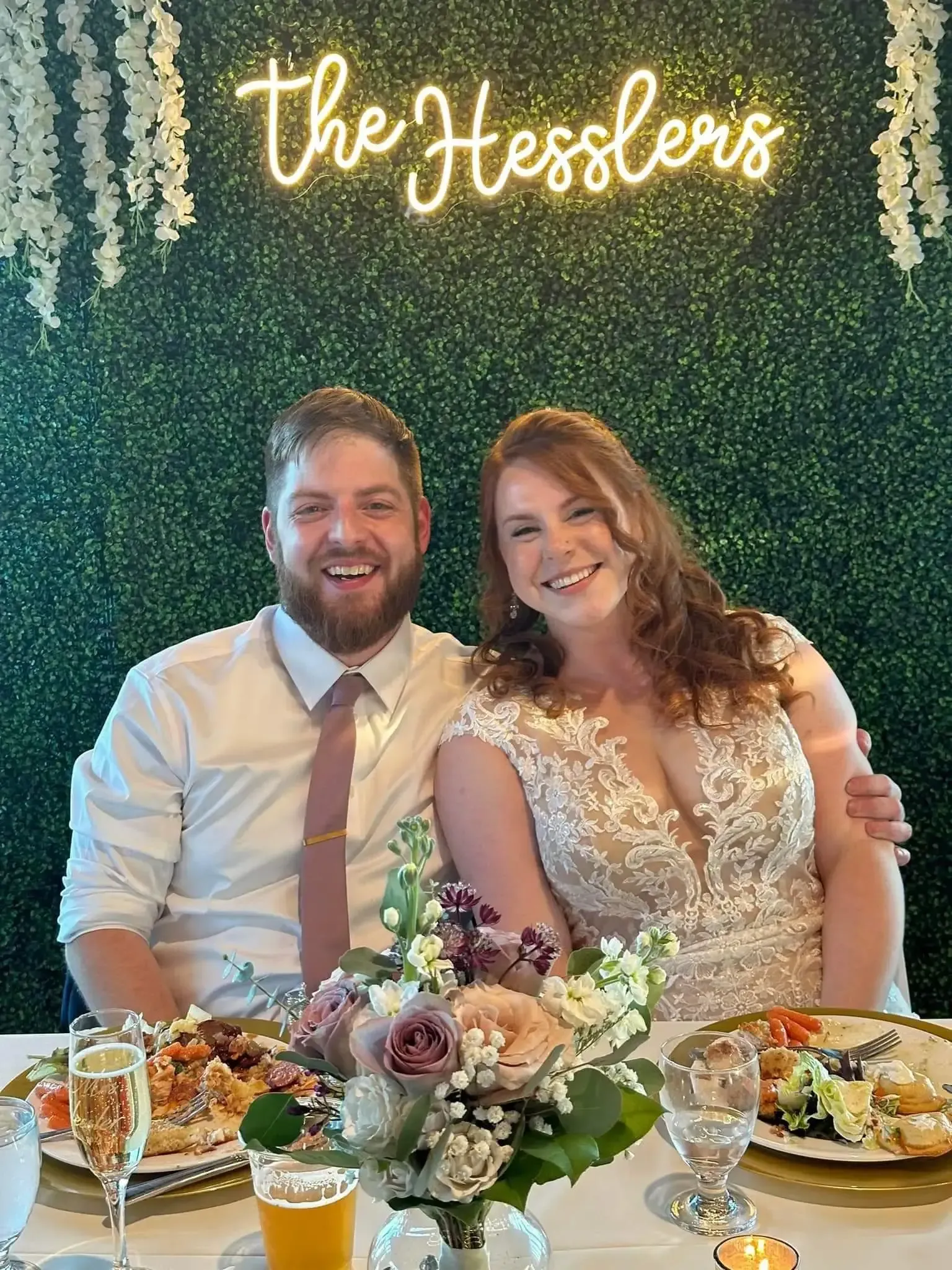 Newlyweds at reception table; couple smiles, floral backdrop and 