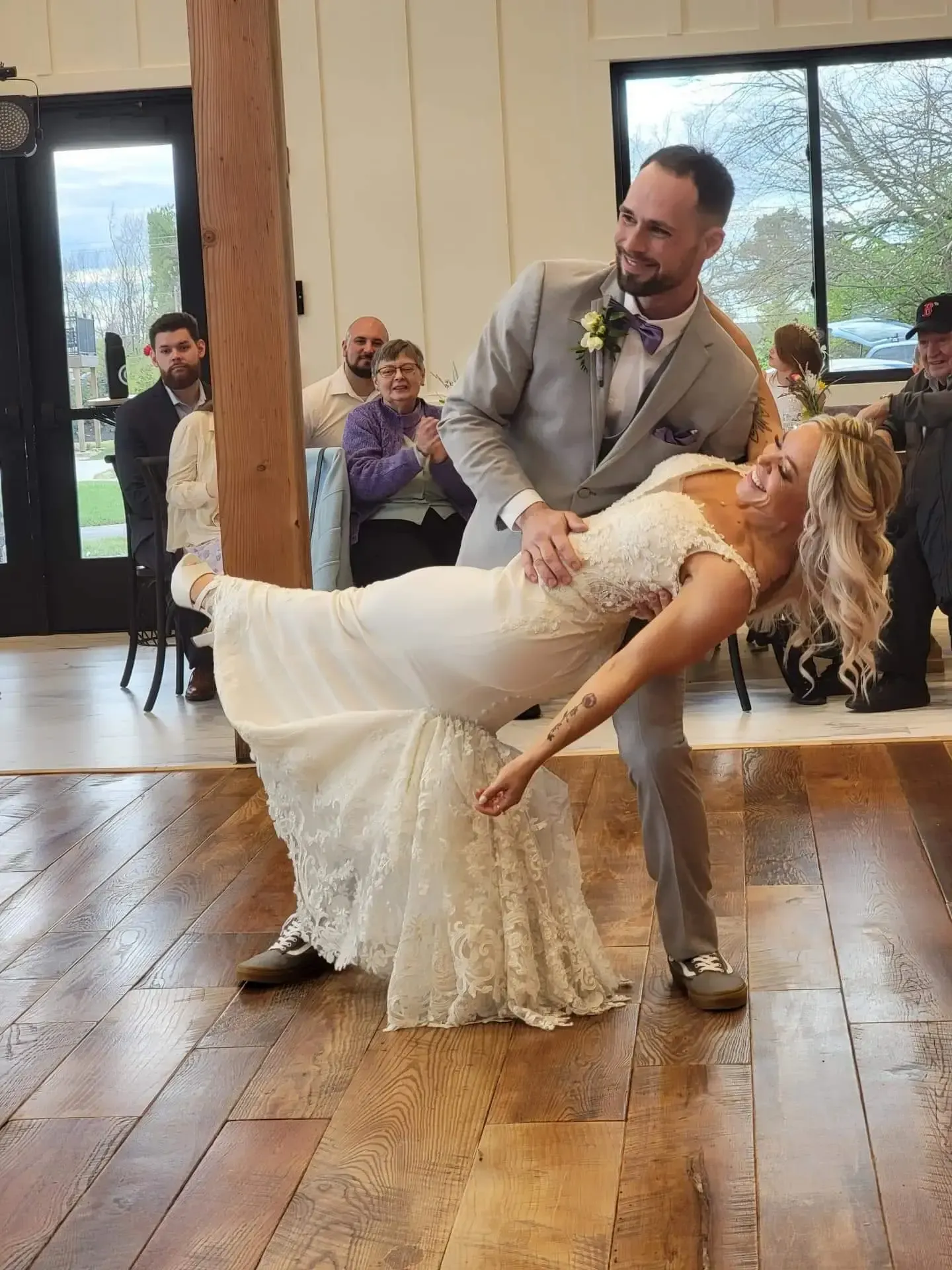 Bride and groom dance at reception. He holds her while she leans back in dress. Wooden floor.
