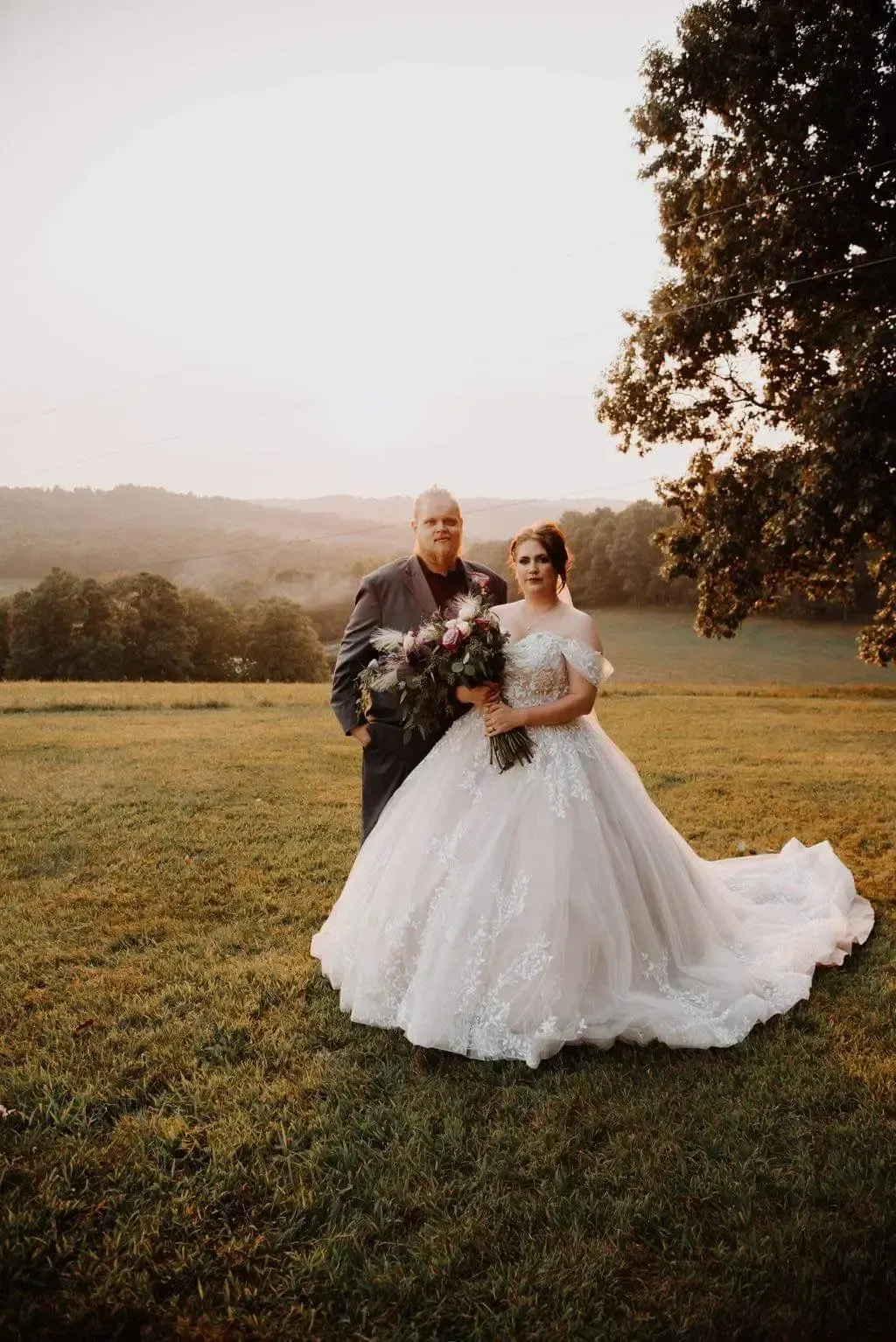 Bride and groom pose on grassy hill at sunset; bride in gown, groom in suit, both holding bouquet.