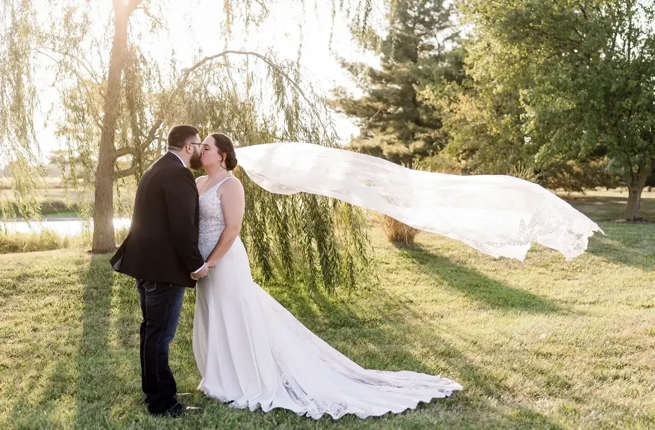 Couple embracing, woman's veil blowing in the wind. Sunny outdoor setting with trees and greenery.