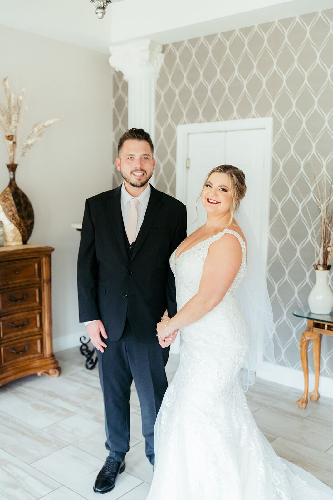 Bride and groom standing indoors, holding hands. The bride wears a white gown, the groom a black suit.