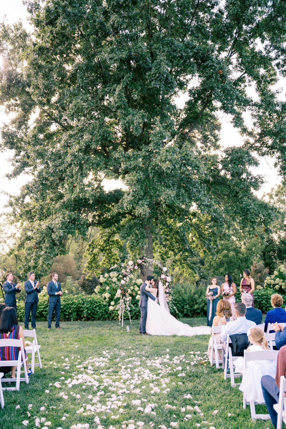 Wedding ceremony under a large tree. Bride and groom stand at altar, guests seated, bridesmaids and groomsmen to the sides.