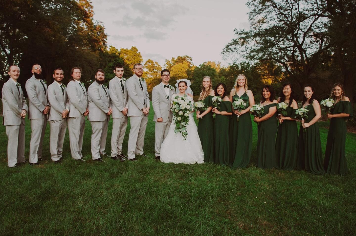 Wedding party: bride in white, bridesmaids in green, groomsmen in gray standing on grass in front of trees.
