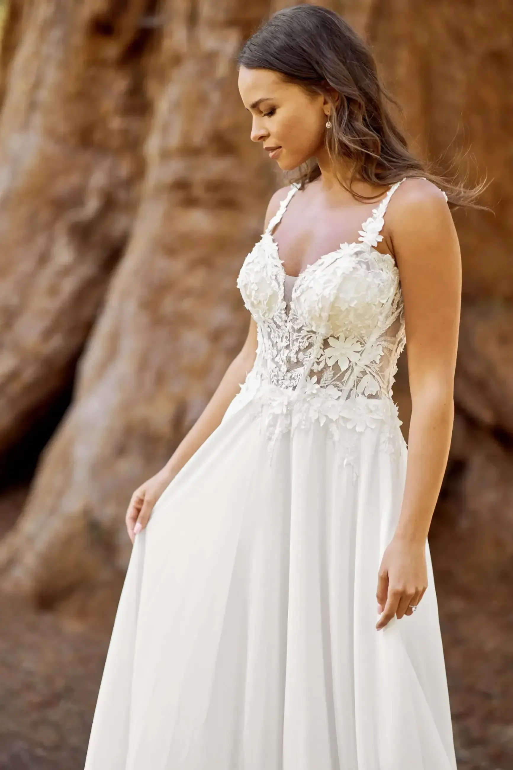 Woman in white wedding dress, outdoors by a large tree, looking down.