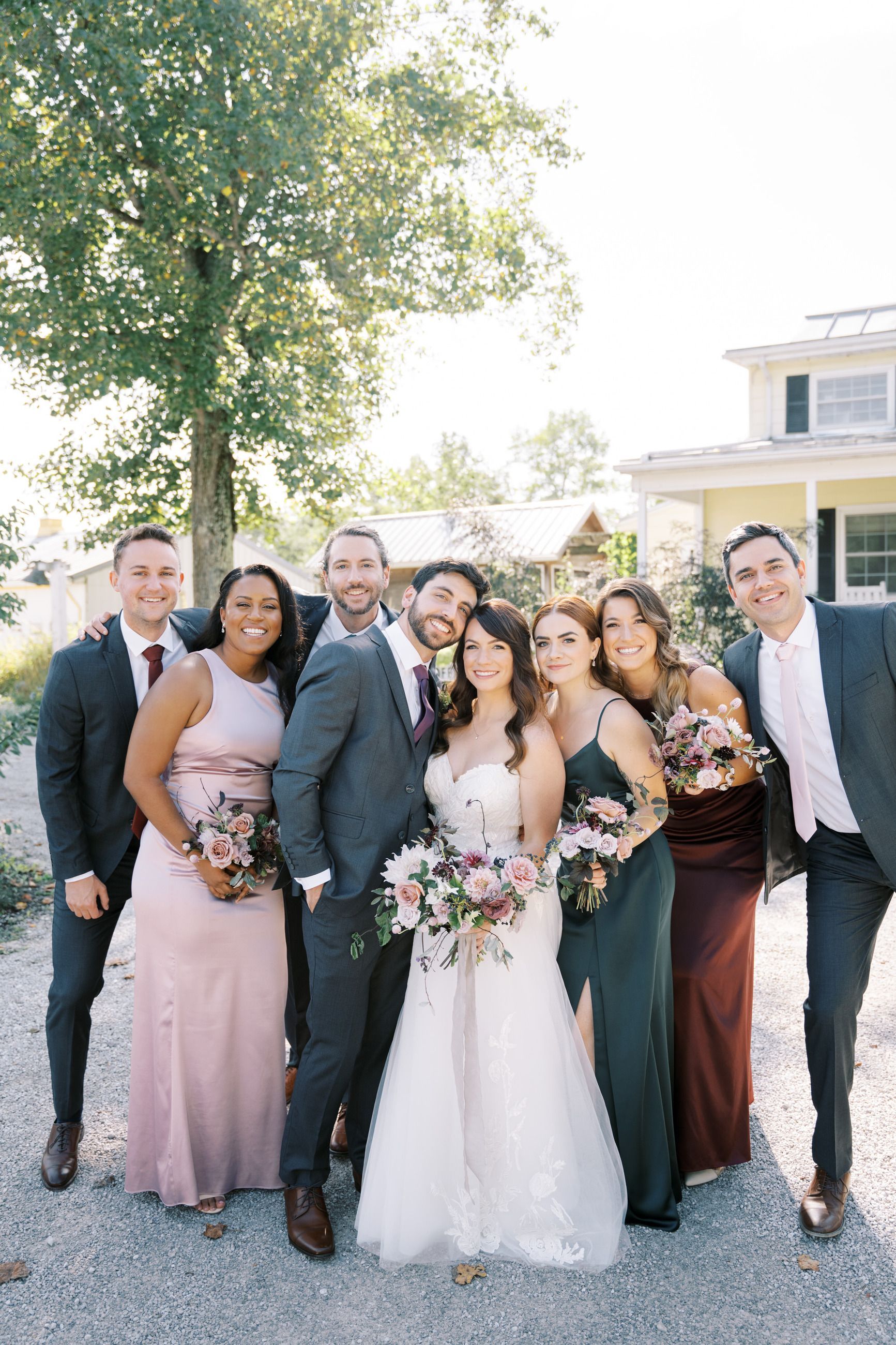 Wedding party smiles outdoors: couple with bridesmaids in various colored gowns and groomsmen in suits.