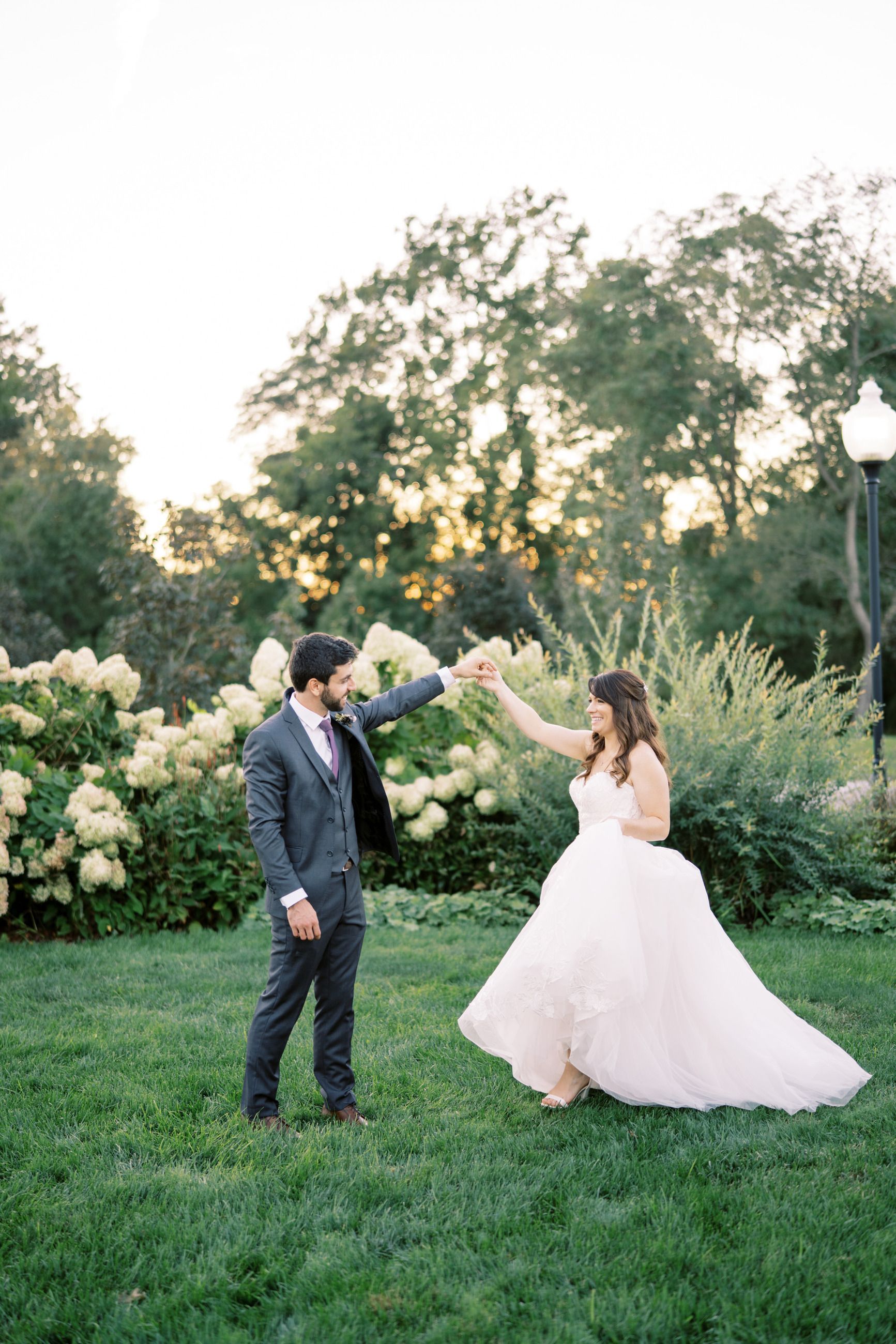 Bride and groom dancing outdoors near white flowers; he in a gray suit, she in a white gown.