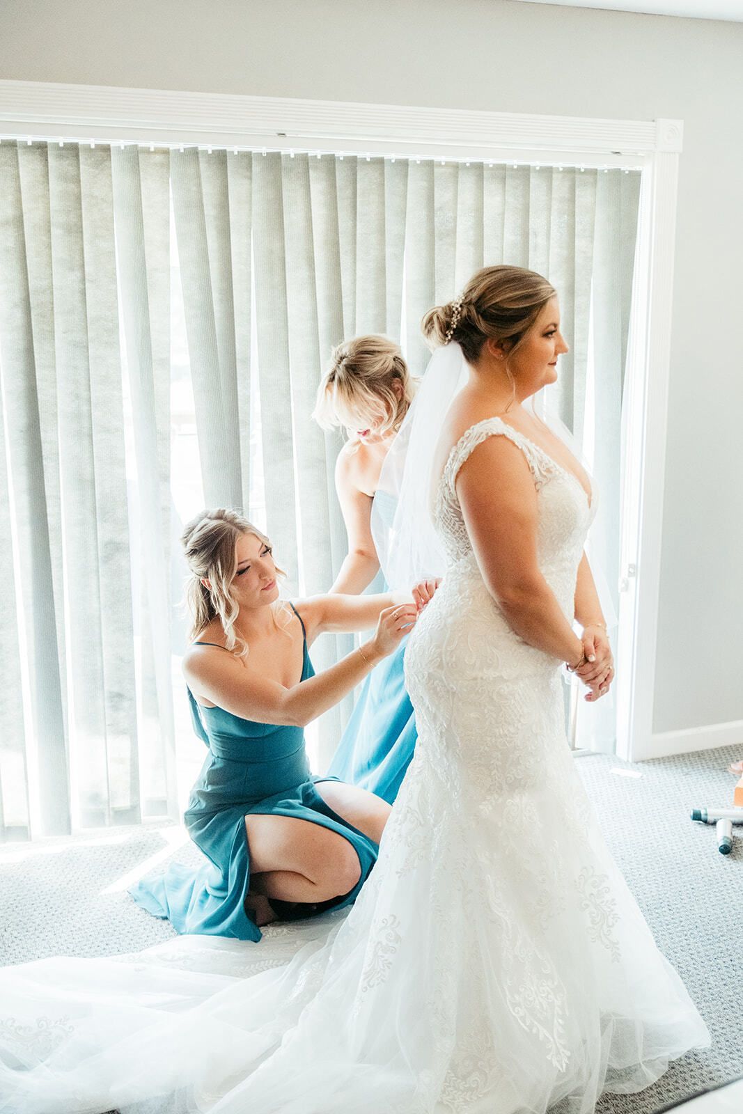 Bride in white dress, assisted by two women in teal dresses, in a room with a large window.