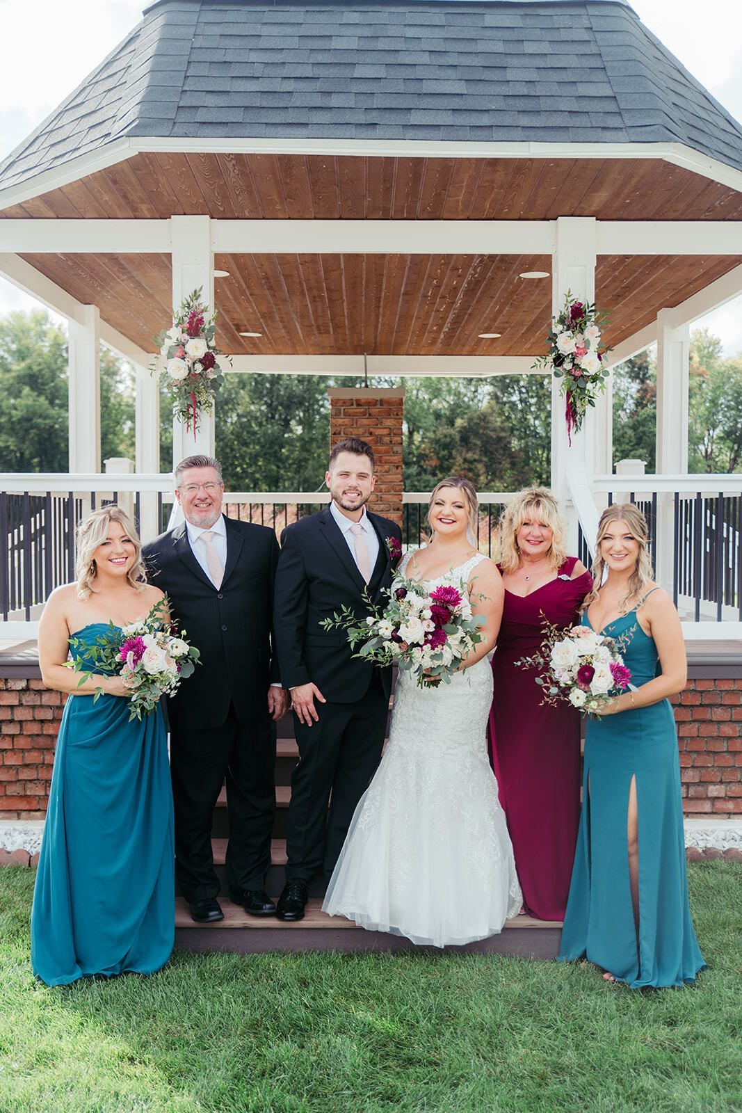 Wedding party posing near a gazebo; bride and groom in the center, flanked by parents and bridesmaids, all holding bouquets.