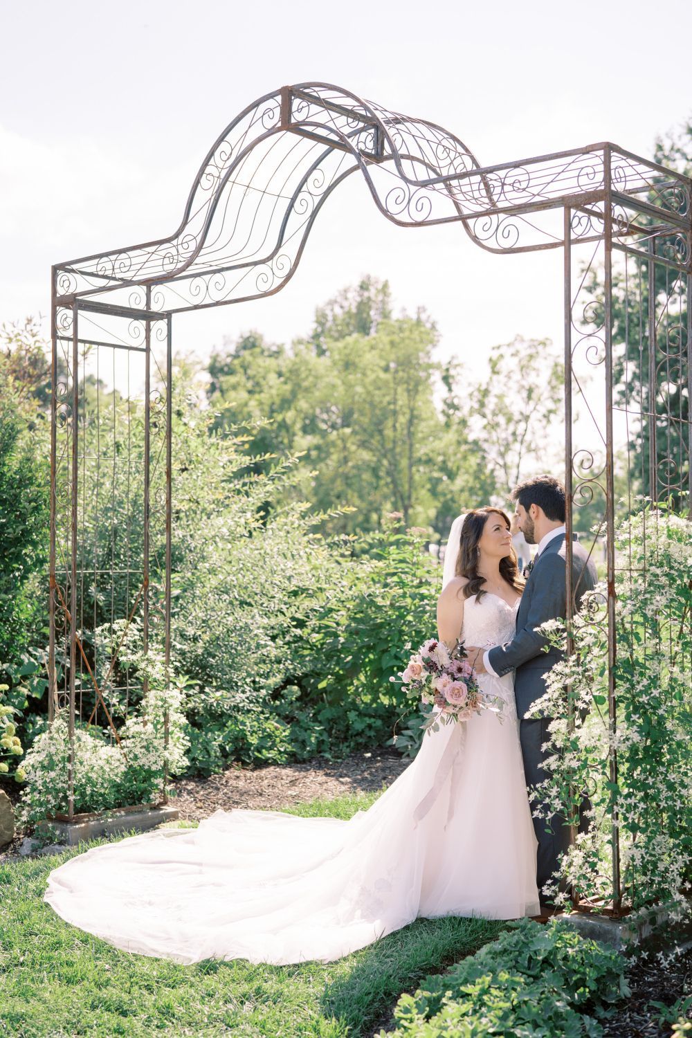 Couple embraces under a wrought iron archway in a lush garden, bride in white gown, holding bouquet.