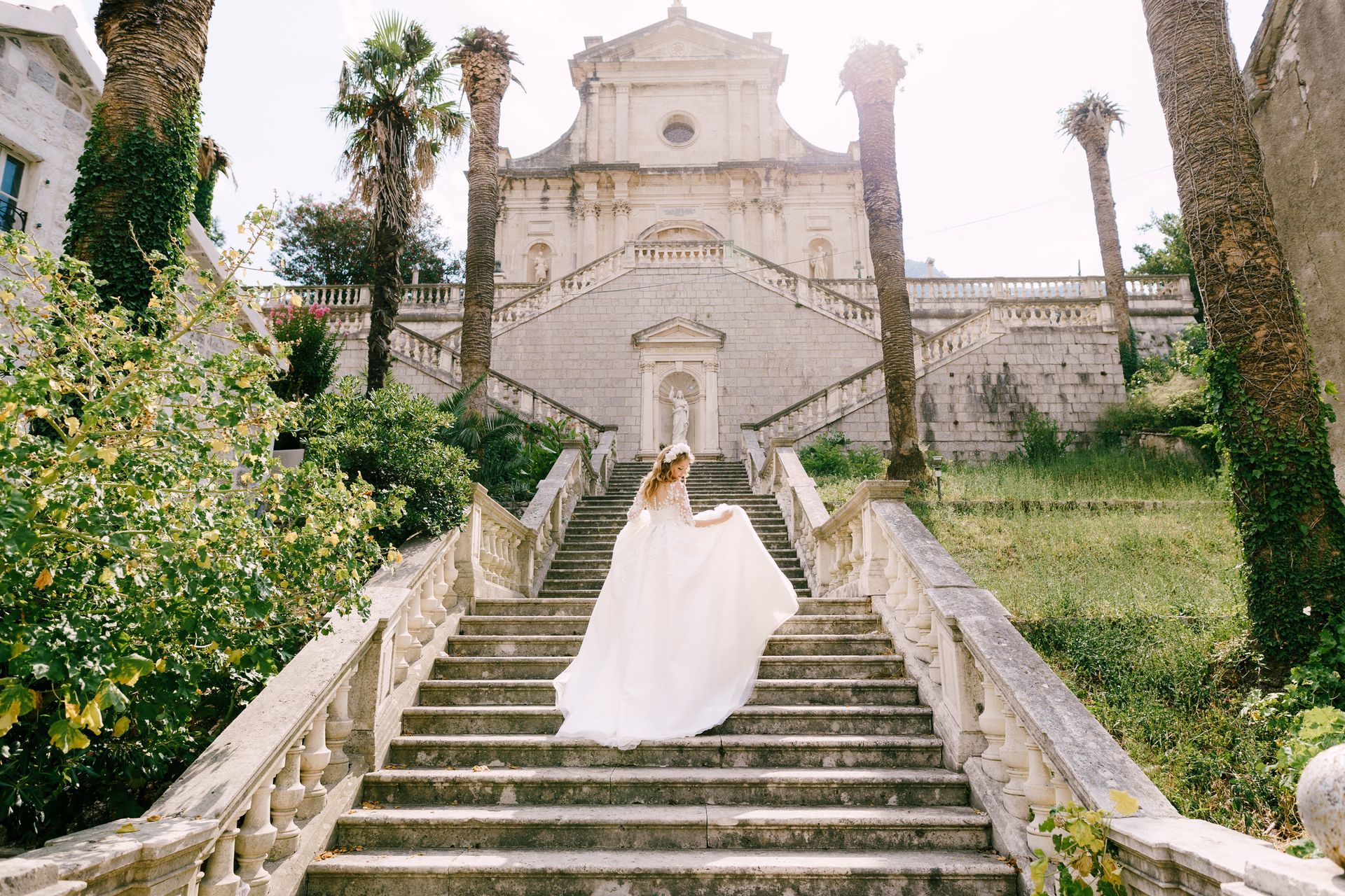 Bride in white dress ascends stone steps toward a church.