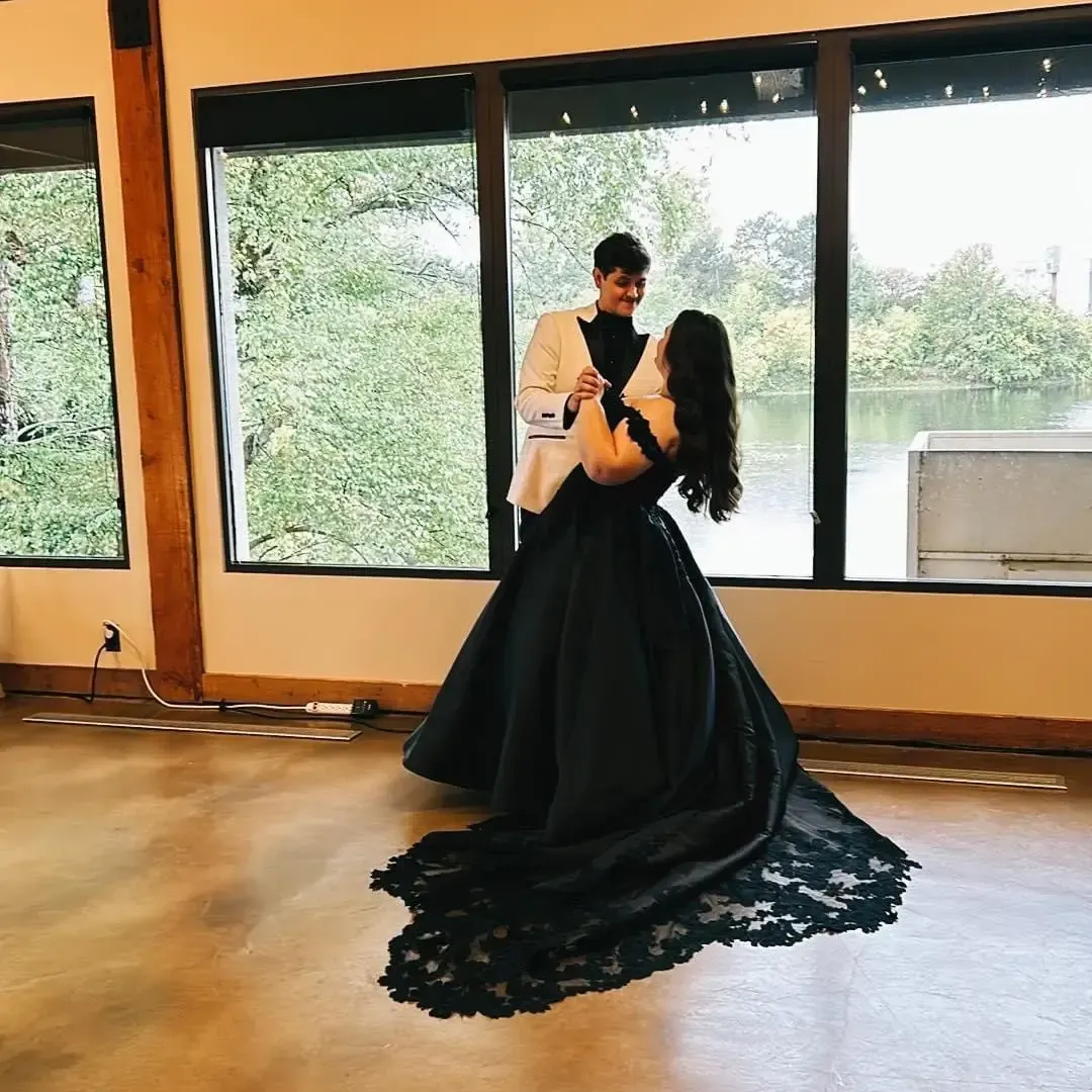 Couple dancing, she in a black gown with a train, he in a white jacket, in front of windows with a lake view.