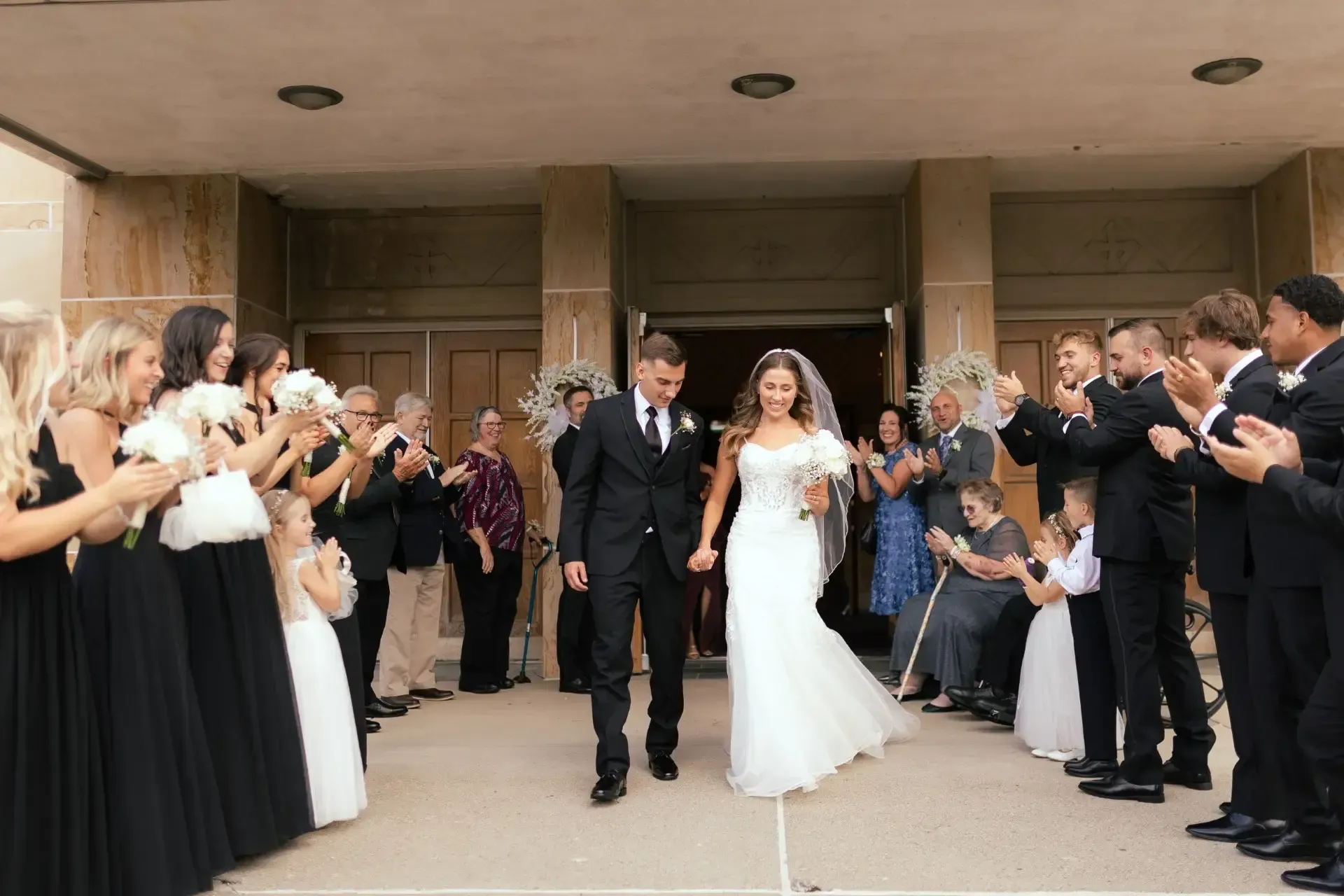 Bride and groom exit church, applauded by wedding party.