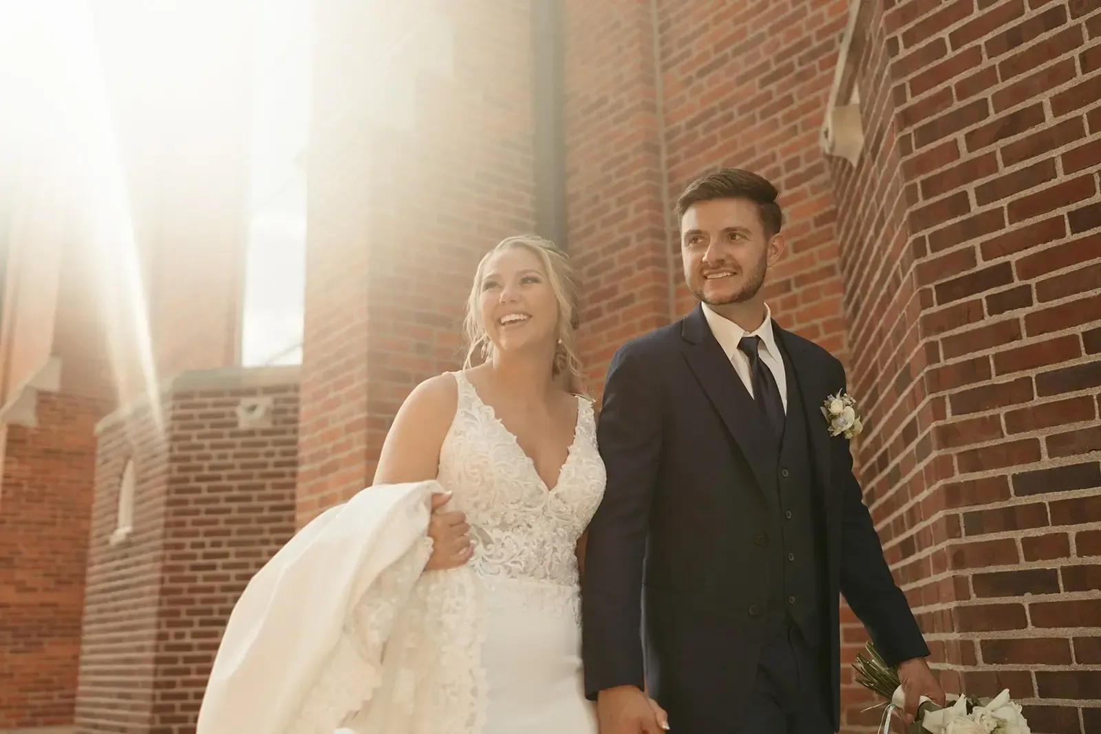 Newlyweds walk, holding hands, near a brick building, sunlight streaming.