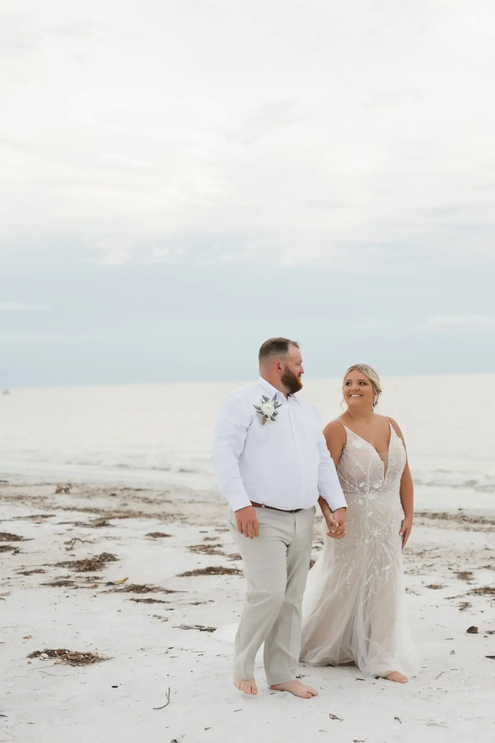 Couple holding hands, smiling at each other on a beach. The bride wears a white dress, the groom a white shirt and khaki pants.