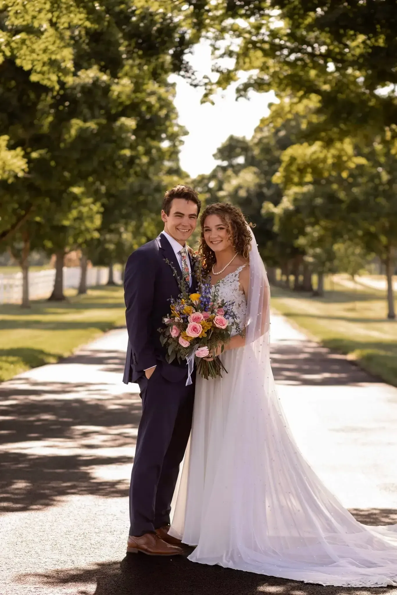 Couple in wedding attire smiles, holding bouquet, on a tree-lined path.