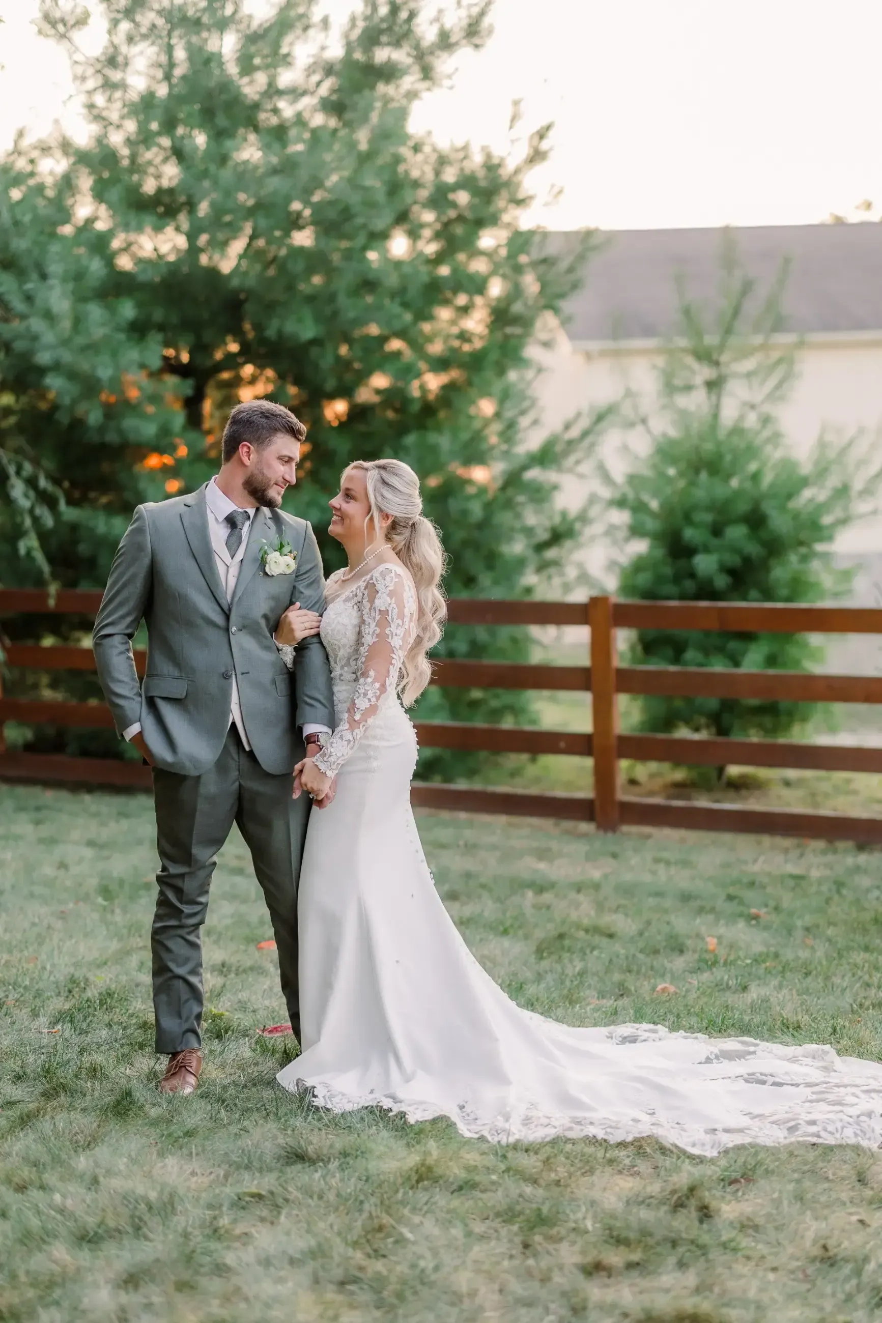 Couple holding hands and smiling, woman in white wedding dress, man in green suit, outdoor setting.