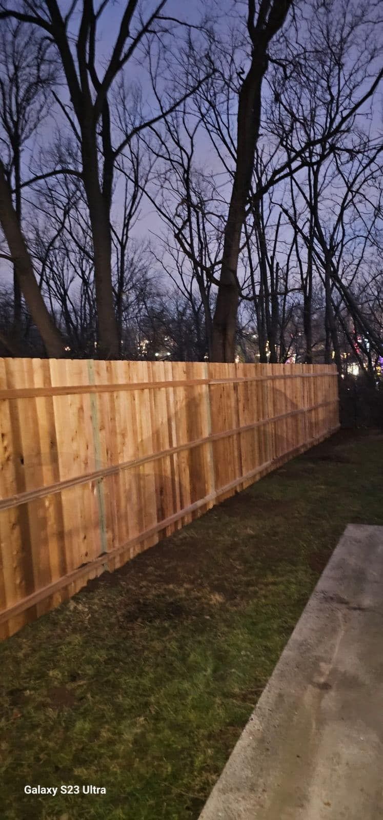 Wooden fence in a backyard, with a lawn and bare trees in the background, at dusk.