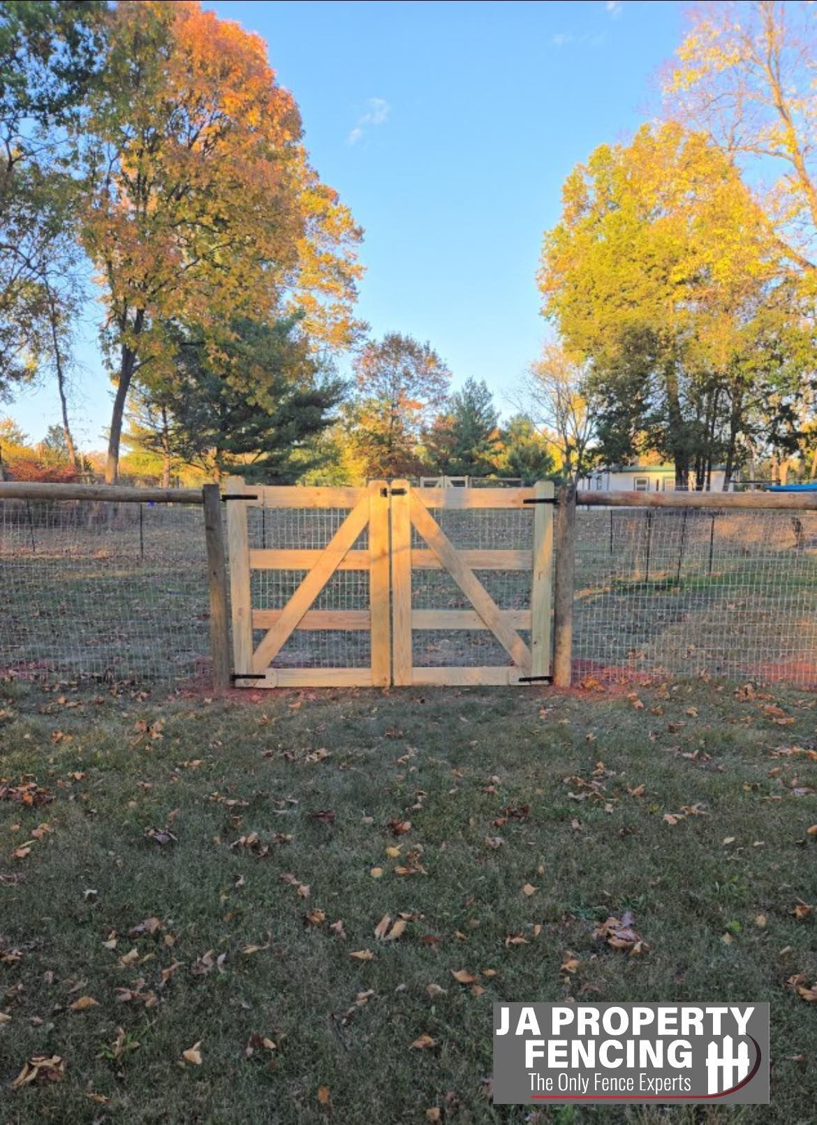 Wooden double gate in a field; wire fence in background. Autumn trees, blue sky. JA Property Fencing logo at bottom.