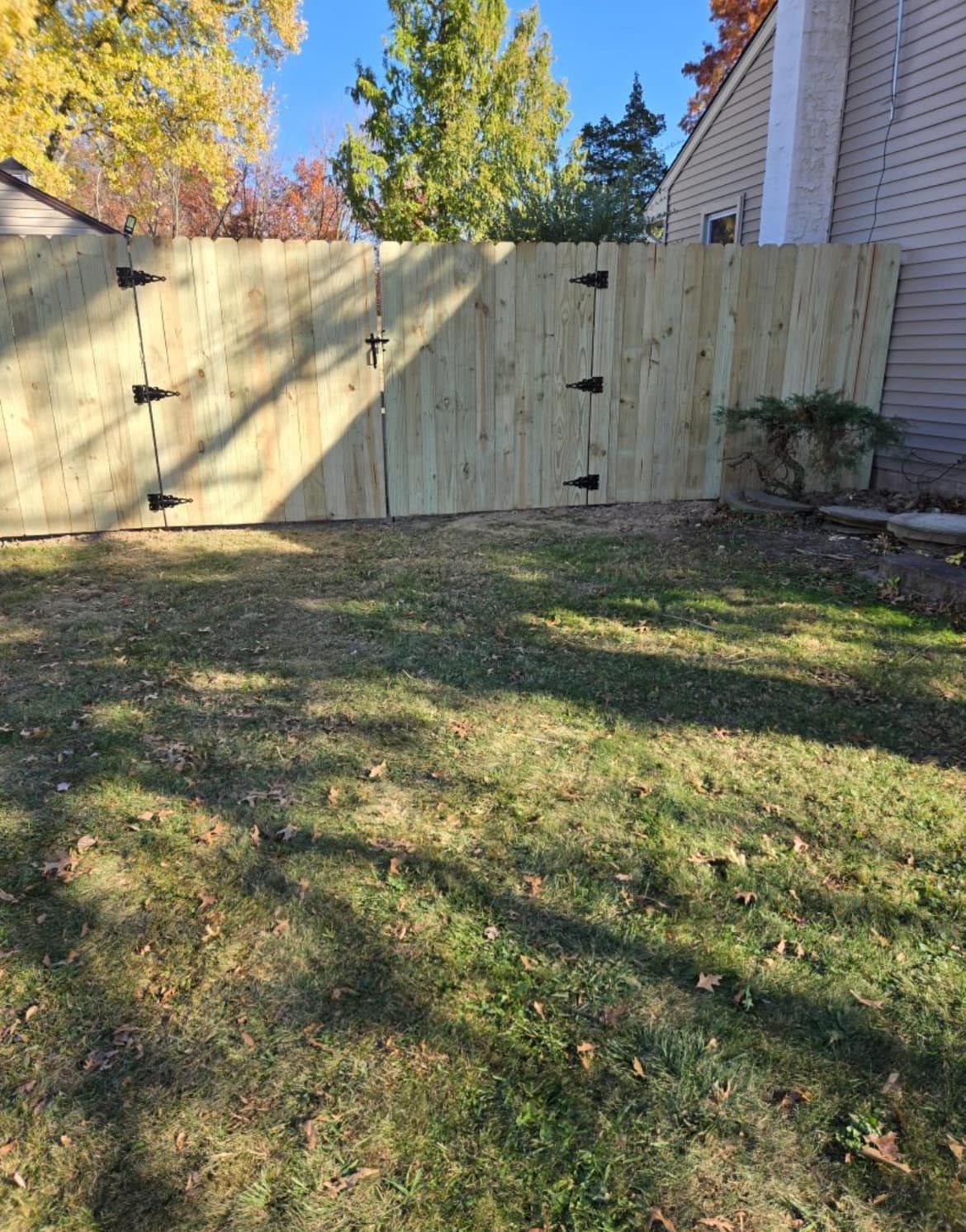 Wooden fence with black hinges in a grassy yard, with a clear sky and trees in the background.