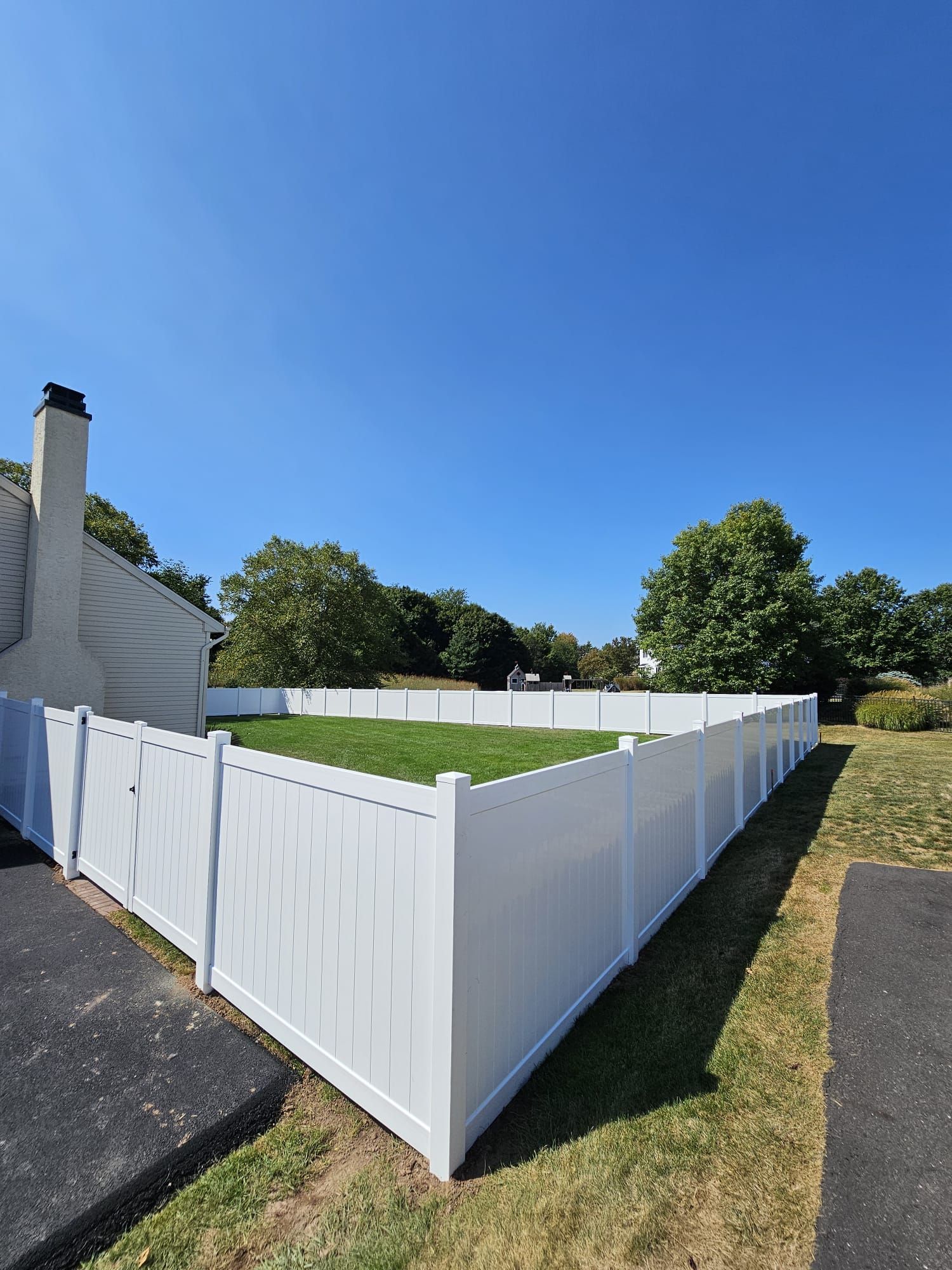 White vinyl fence enclosing a paved area with a basketball hoop. The sun casts shadows.