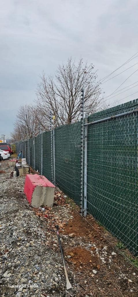 A green privacy fence topped with barbed wire runs along a gravel and dirt area under a cloudy sky.