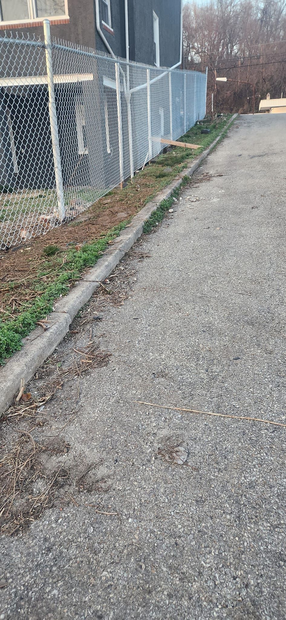 Gravel driveway next to a chain link fence and small strip of grass. Gray building in the background.