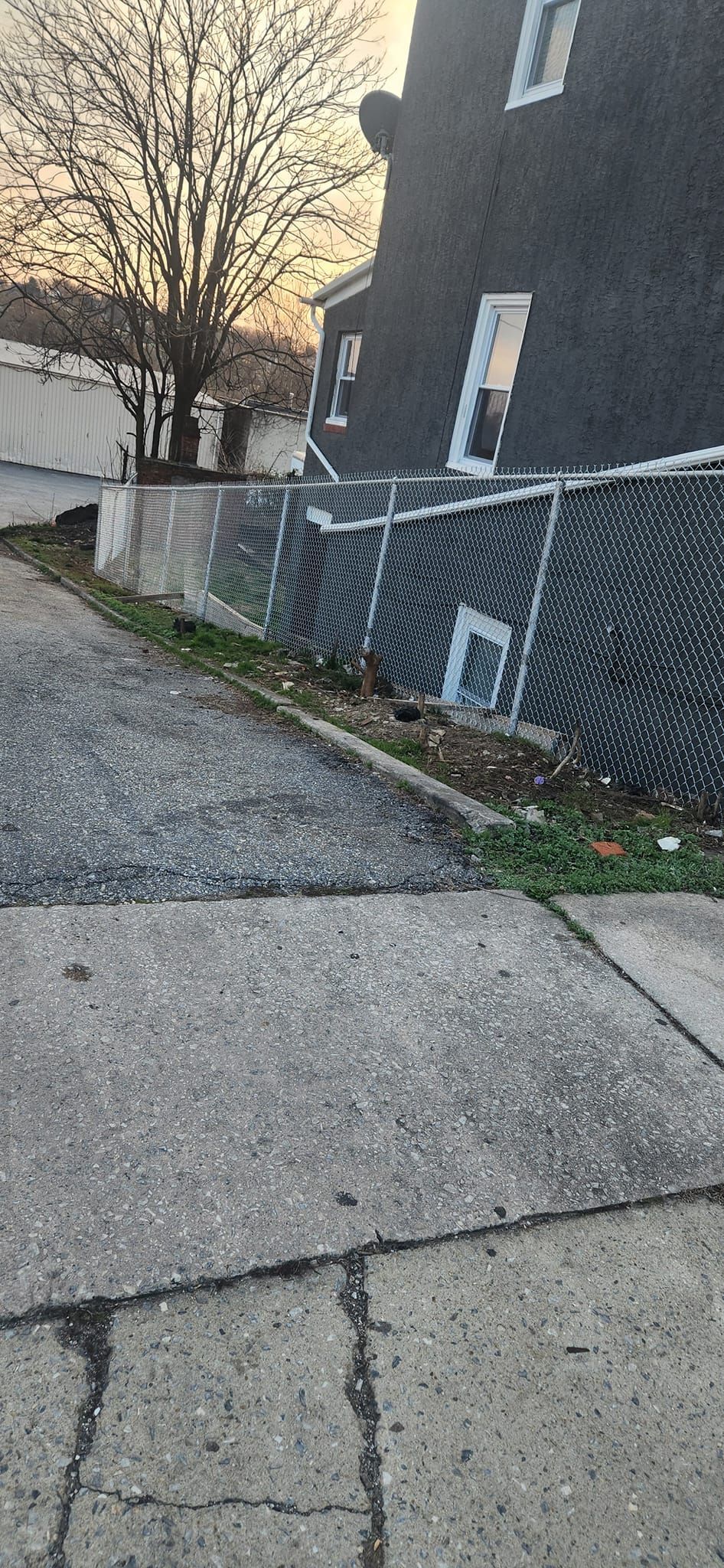 A concrete sidewalk and driveway lead past a chain-link fence and a dark-colored building.