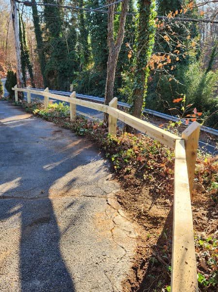 Wooden fence along a paved path, with a tree and foliage behind it.