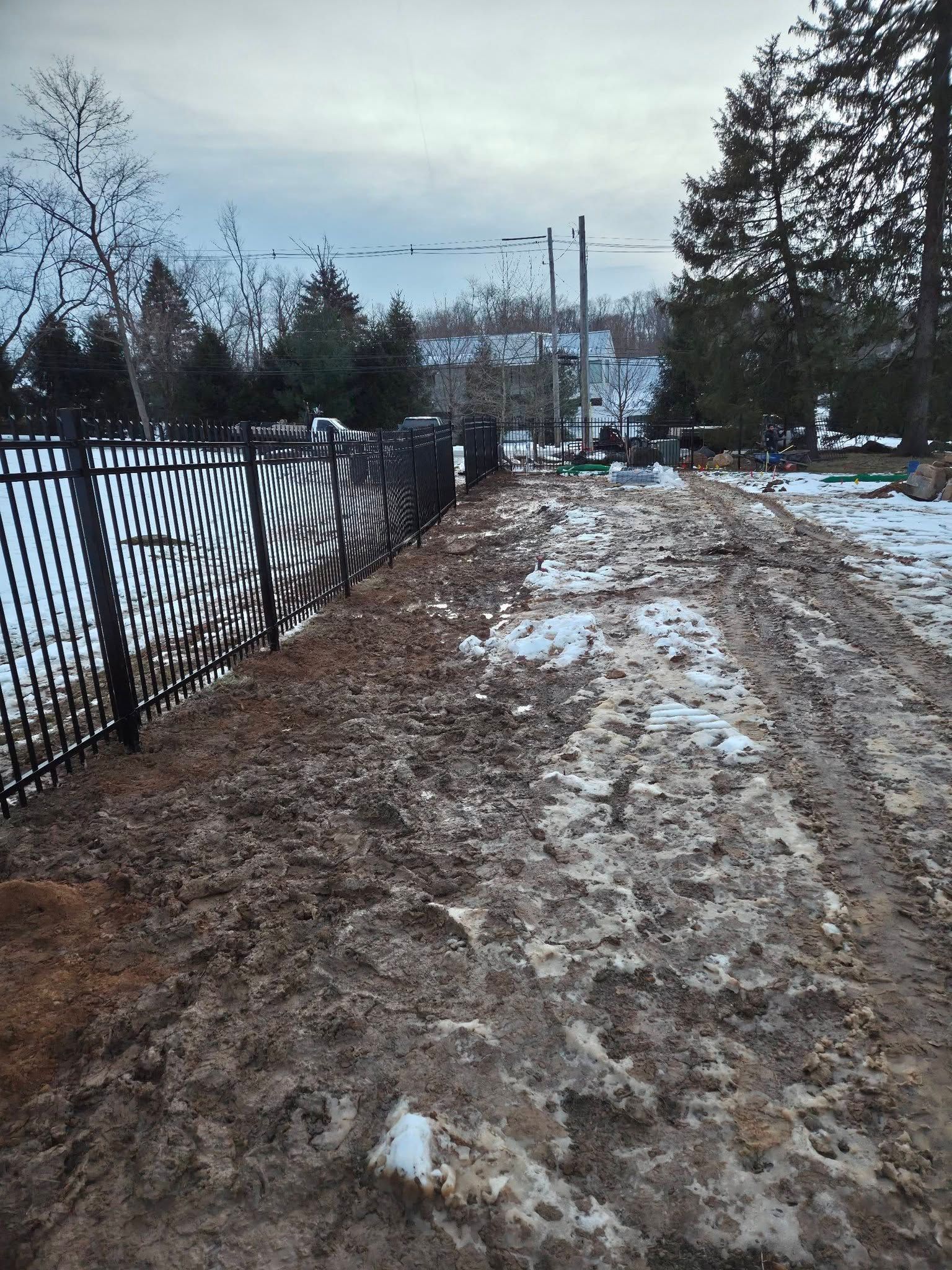 Muddy driveway next to black metal fence. Some snow present. Cloudy sky, trees in the background.
