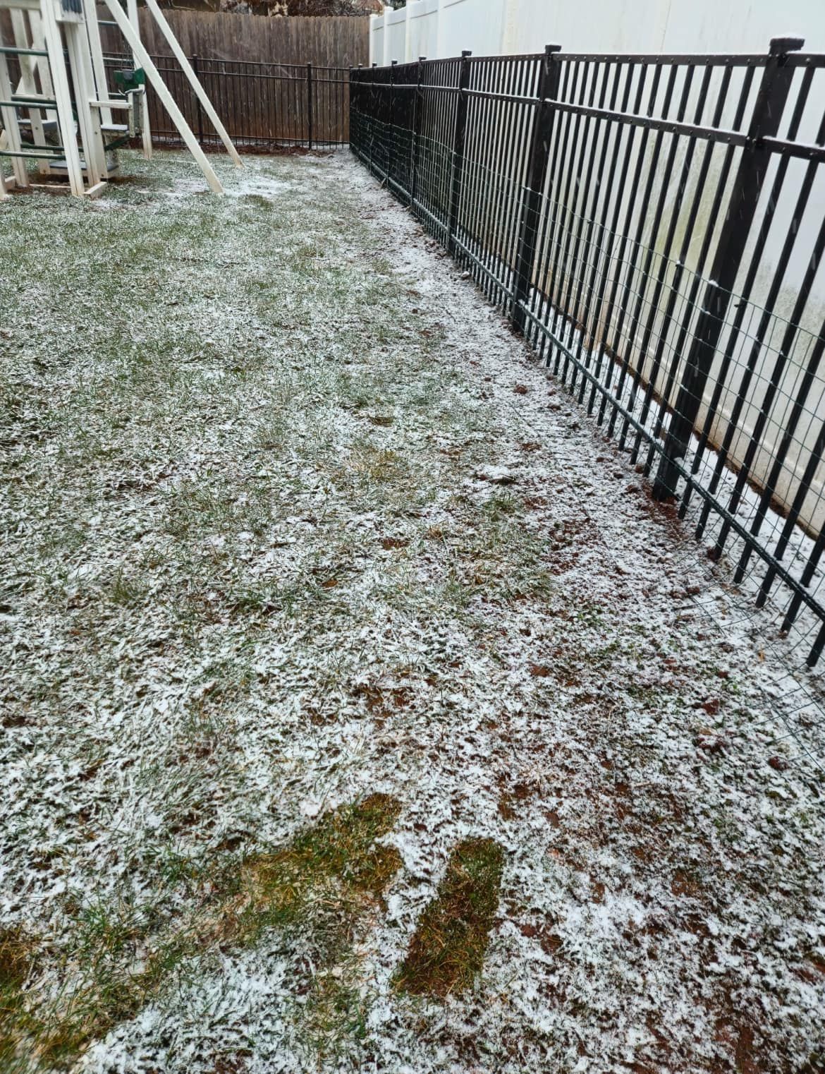 Backyard lawn with patchy snow, black metal fence, and a swing set.