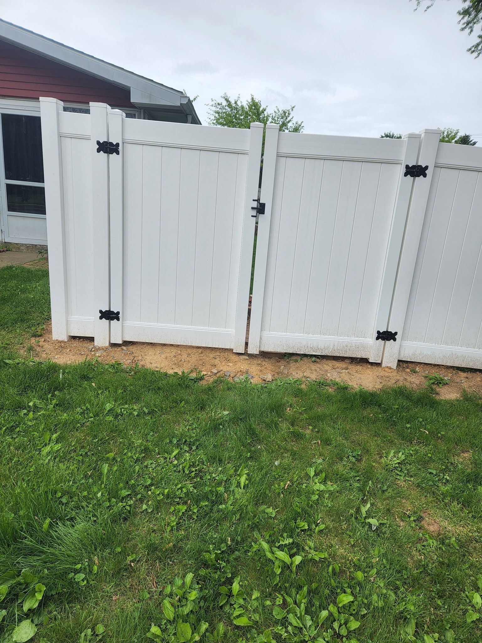 White vinyl fence with gate, set in dirt and grass, against a building with a red roof.