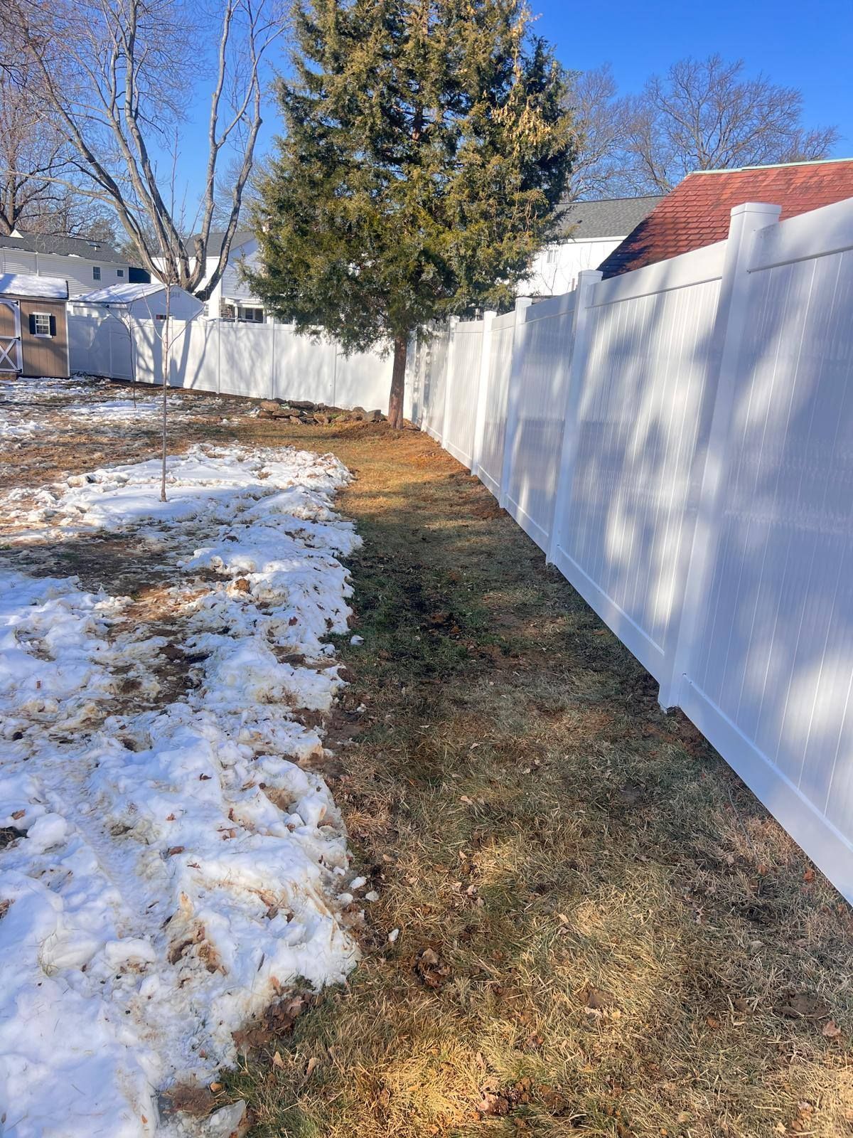 Backyard with white fence, patches of snow, and brown grass. A tree stands near the fence.