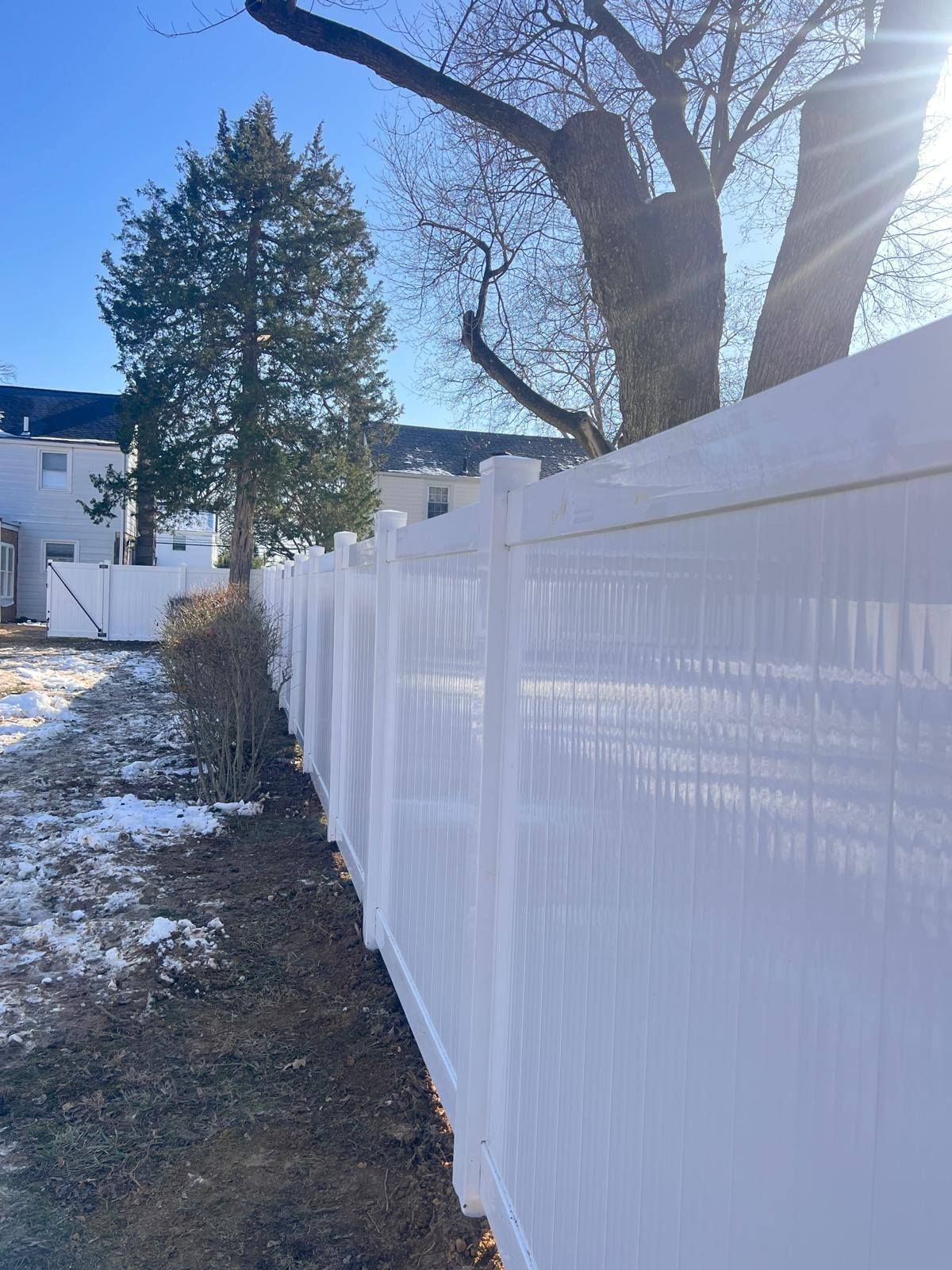 White vinyl fence in a yard on a sunny day, with a small patch of snow and bare trees.