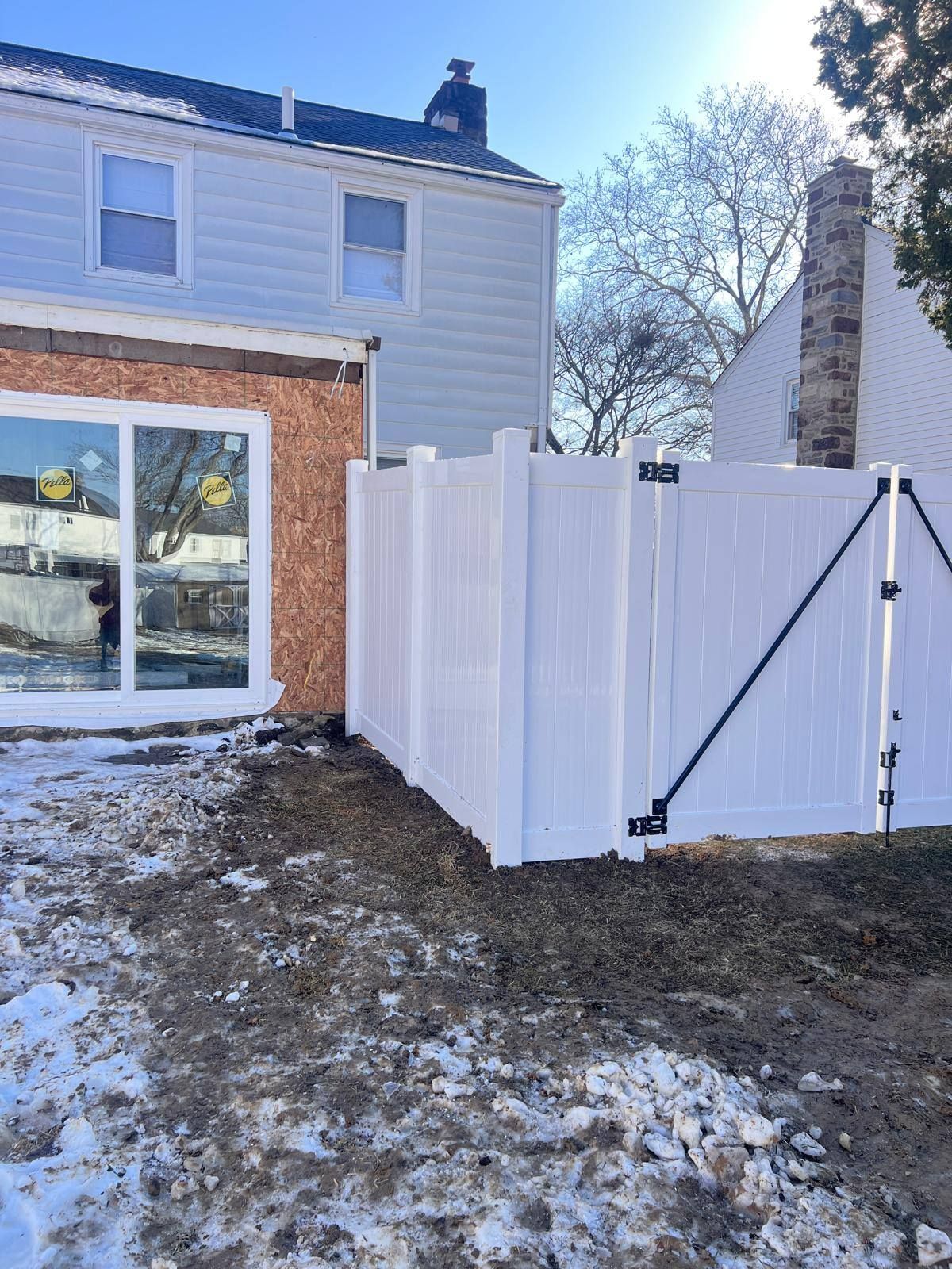 White vinyl fence next to a two-story house with a partially constructed wall. Snow covers the ground.