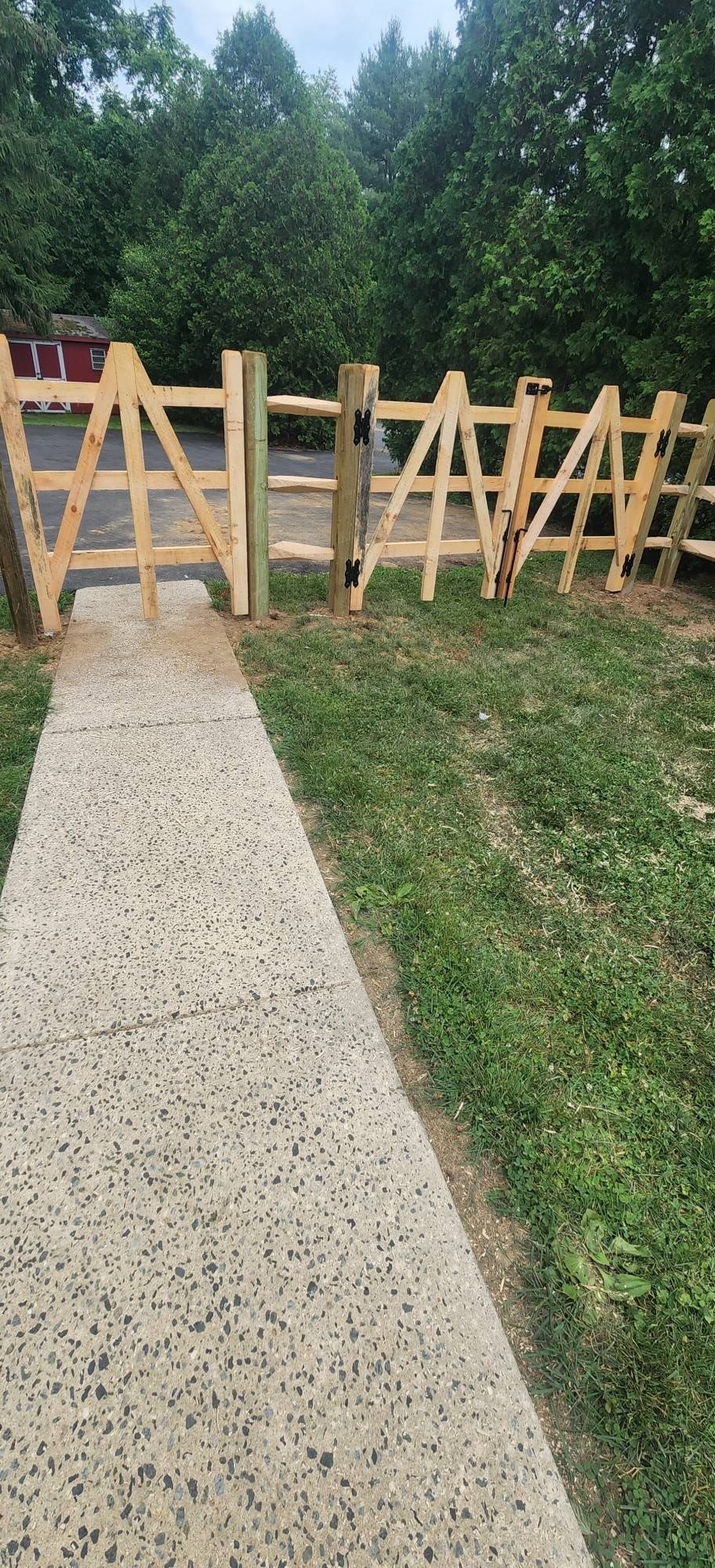 A wooden fence with a gate along a concrete pathway leading into a grassy area, trees in the background.