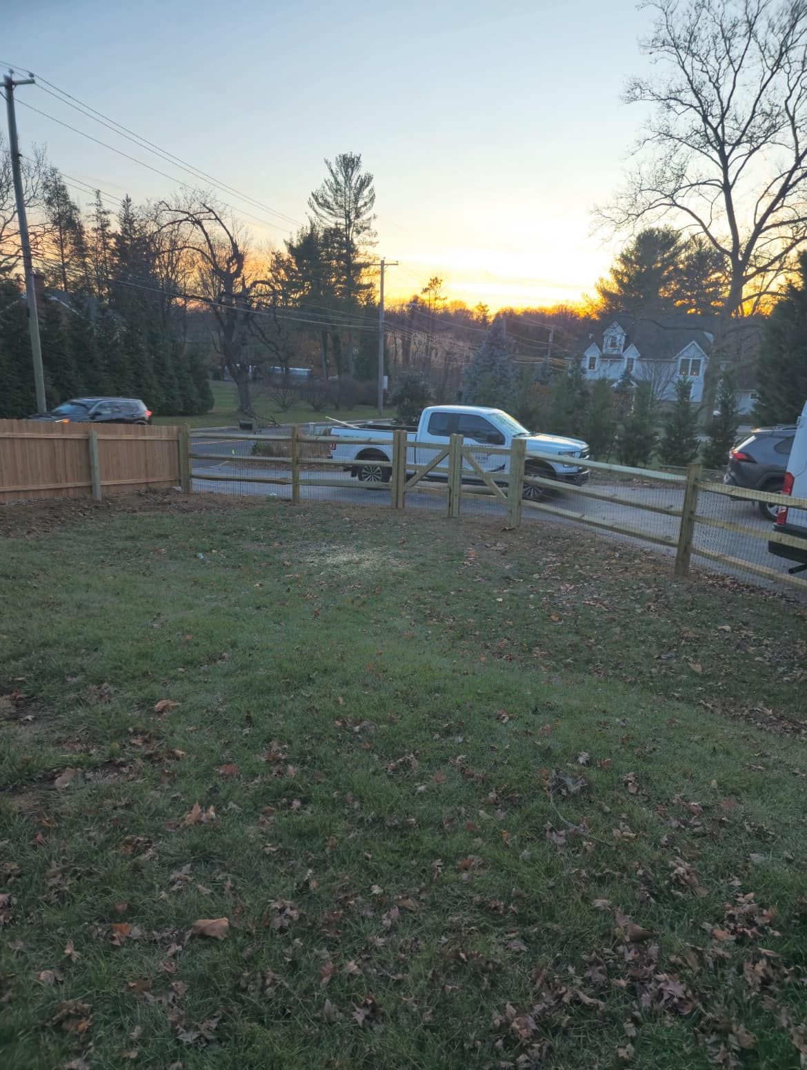 A white pickup truck parked behind a wooden fence, in front of a house, at dusk.
