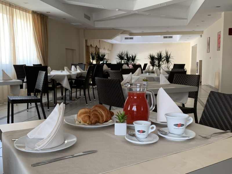 Tavolo per la colazione apparecchiato con croissant, succo di frutta e caffè, in una sala da pranzo luminosa con tavoli e sedie.