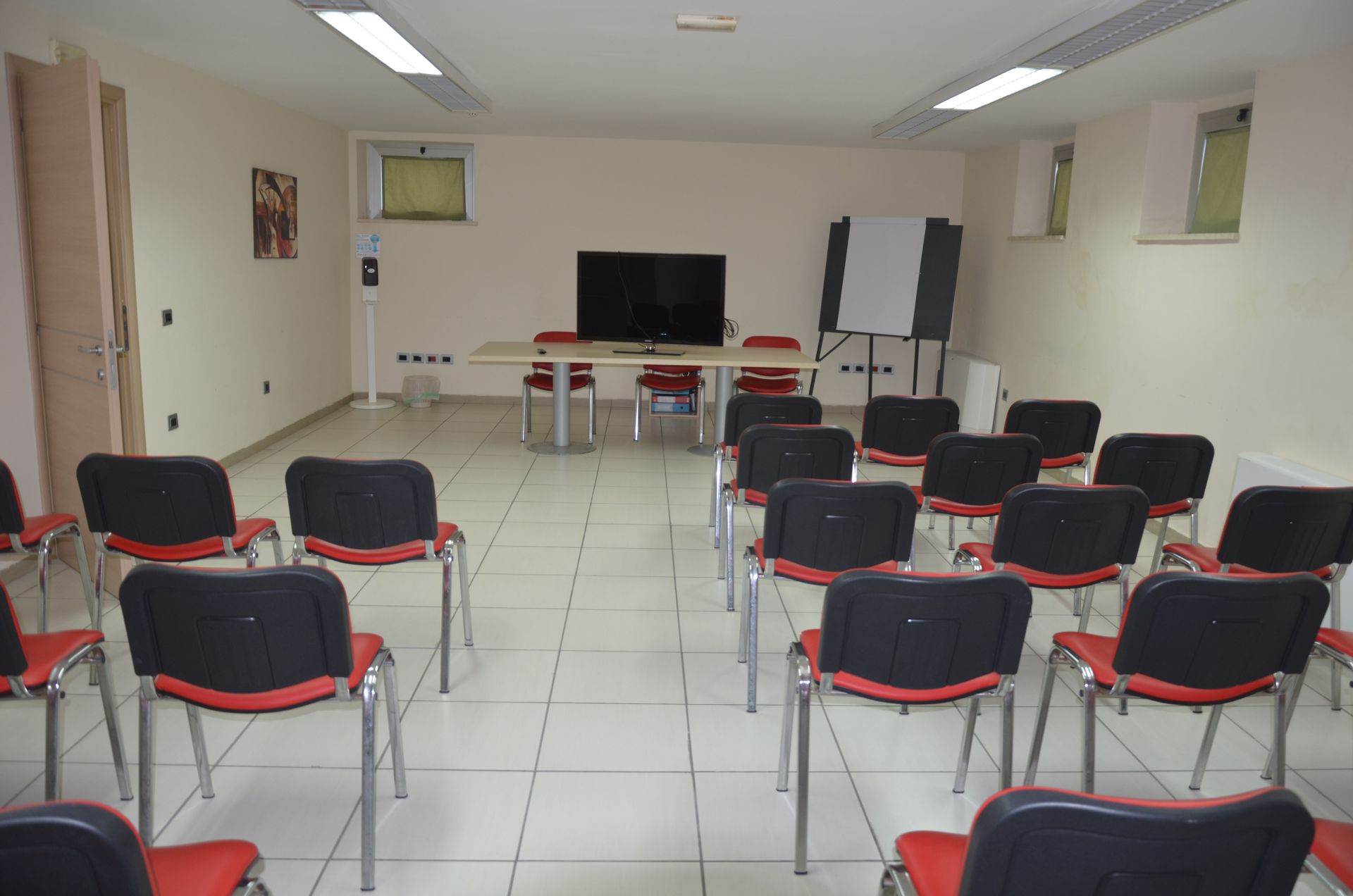Sala conferenze vuota con file di sedie rosse e nere di fronte a un tavolo con TV. Lavagna e tavolo di fronte.