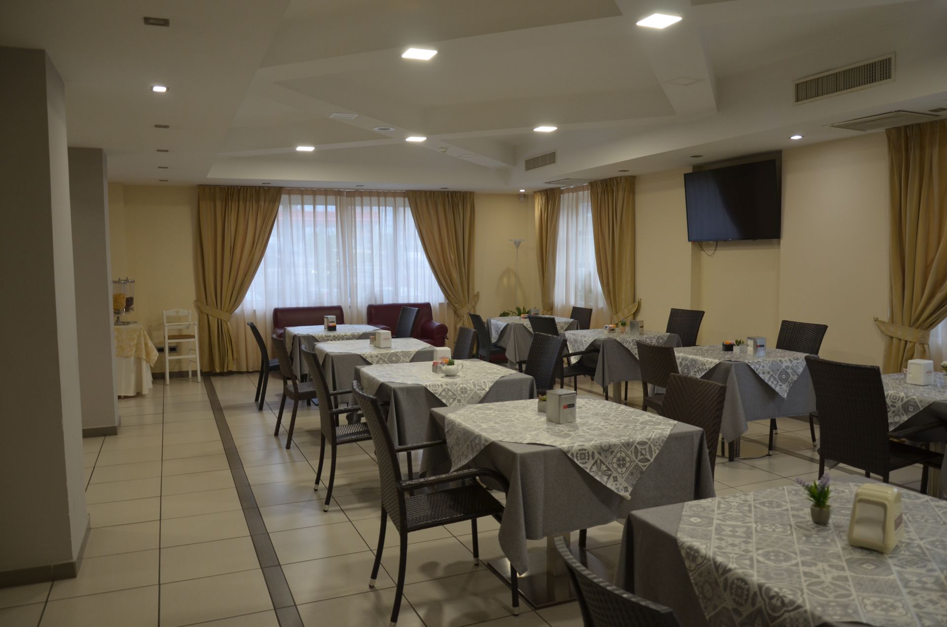 Sala ristorante con tavoli, sedie, tende e TV. La sala è caratterizzata da colori neutri.