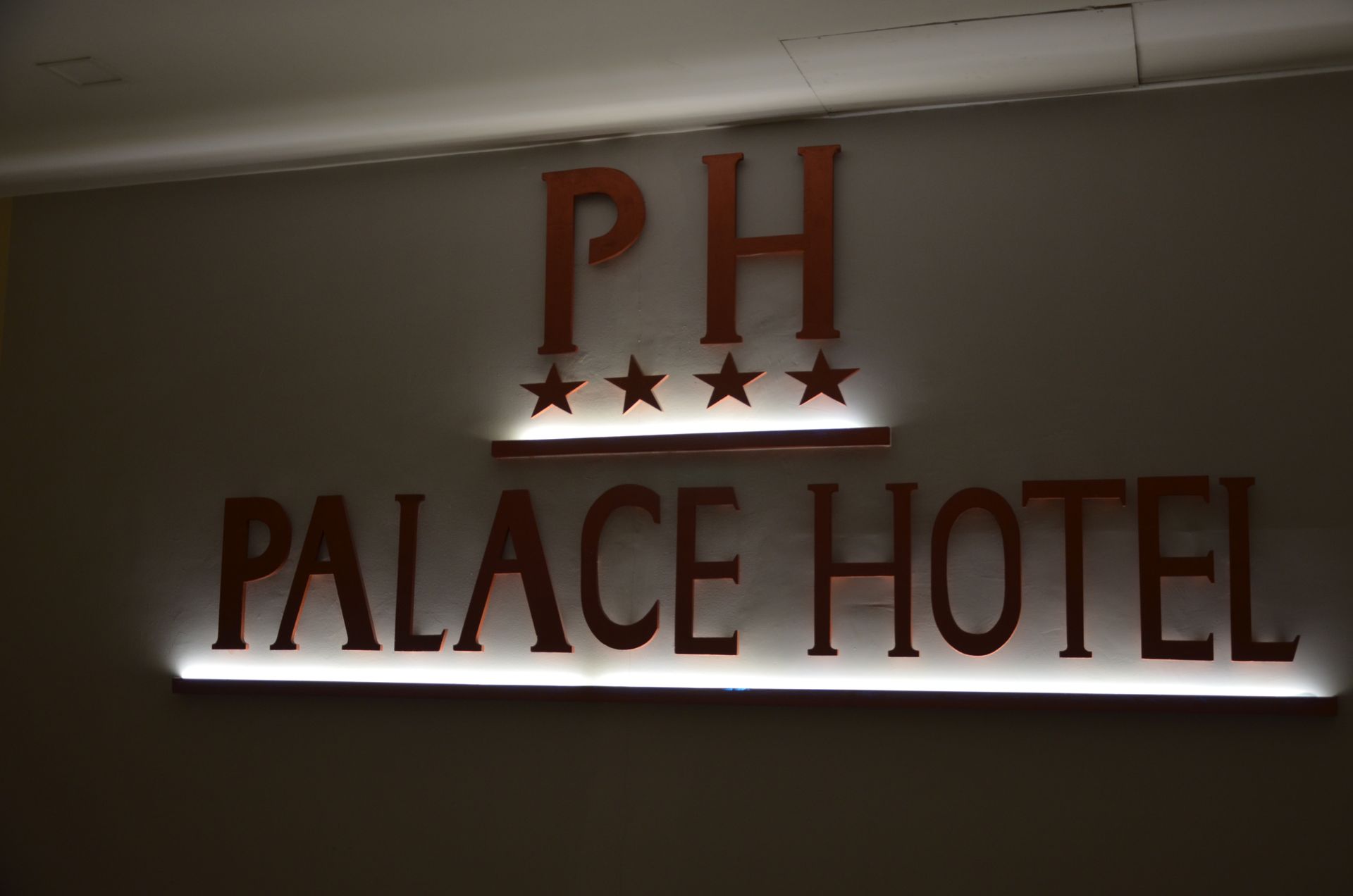 Insegna del PH Palace Hotel con lettere in bronzo e quattro stelle, retroilluminata su una parete grigia.