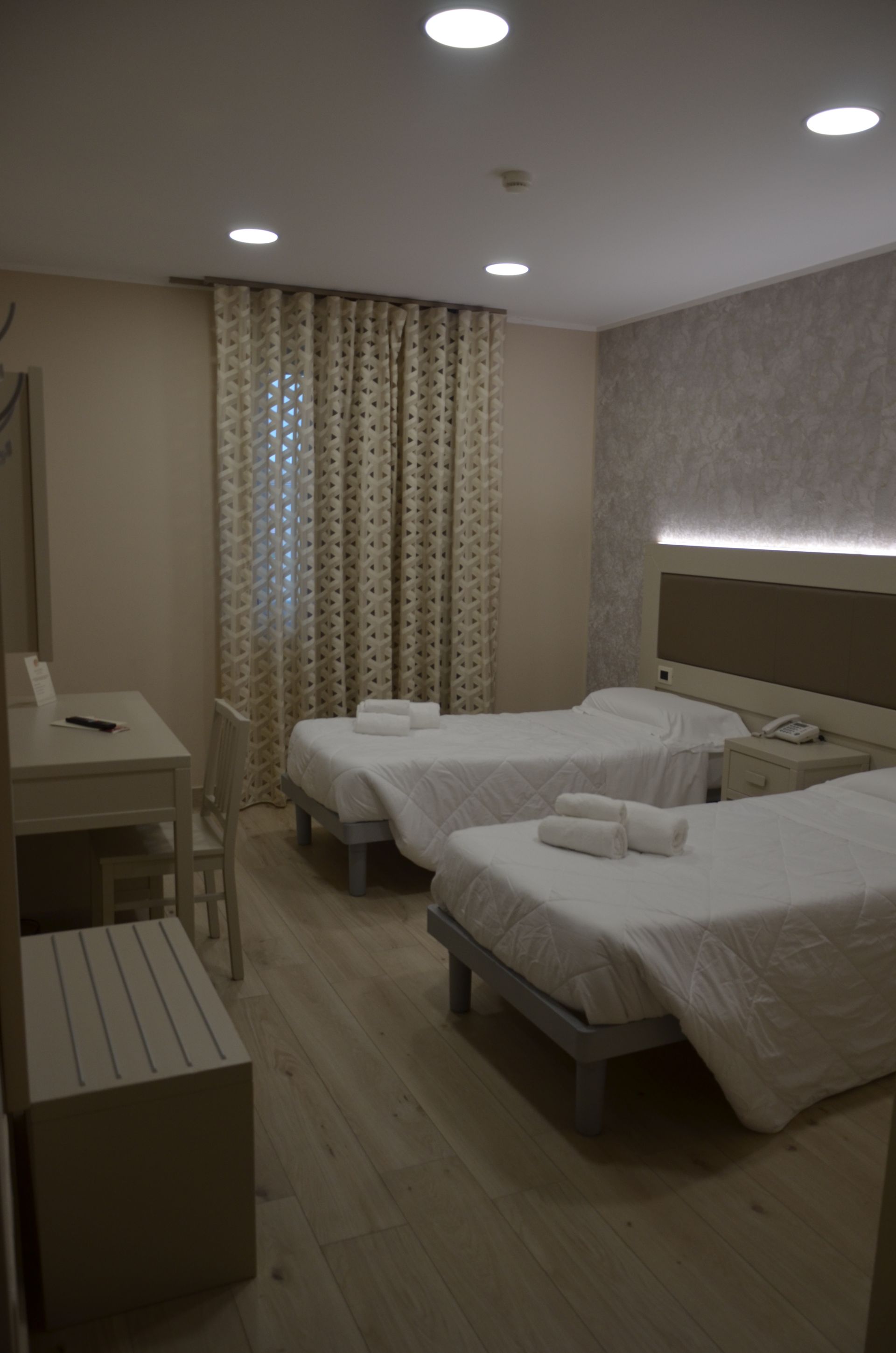 Due letti in una stanza d'albergo, con scrivania, panca e tende a fantasia.