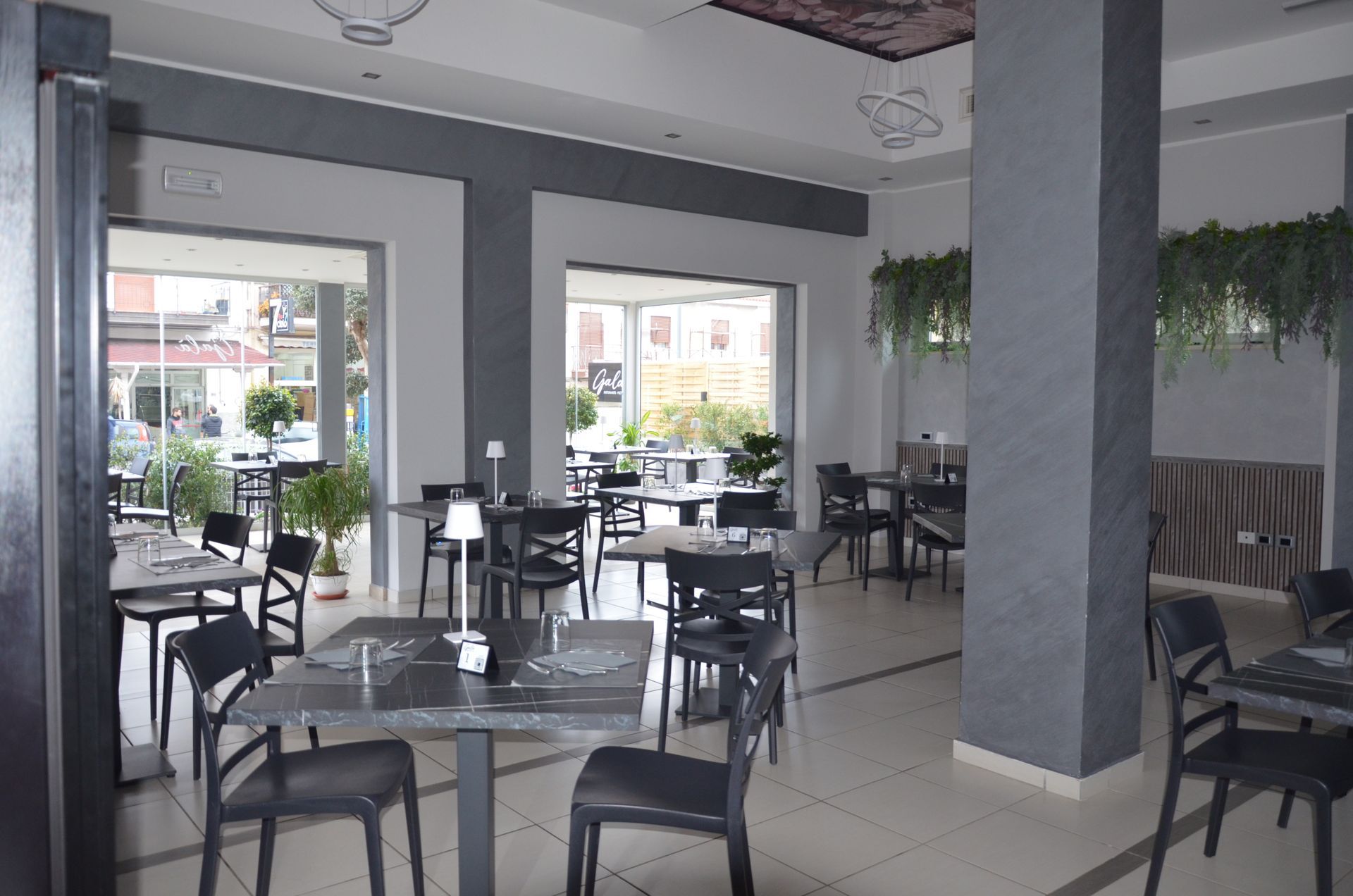 Interno del ristorante con tavoli e sedie, ampie finestre e piante decorative.