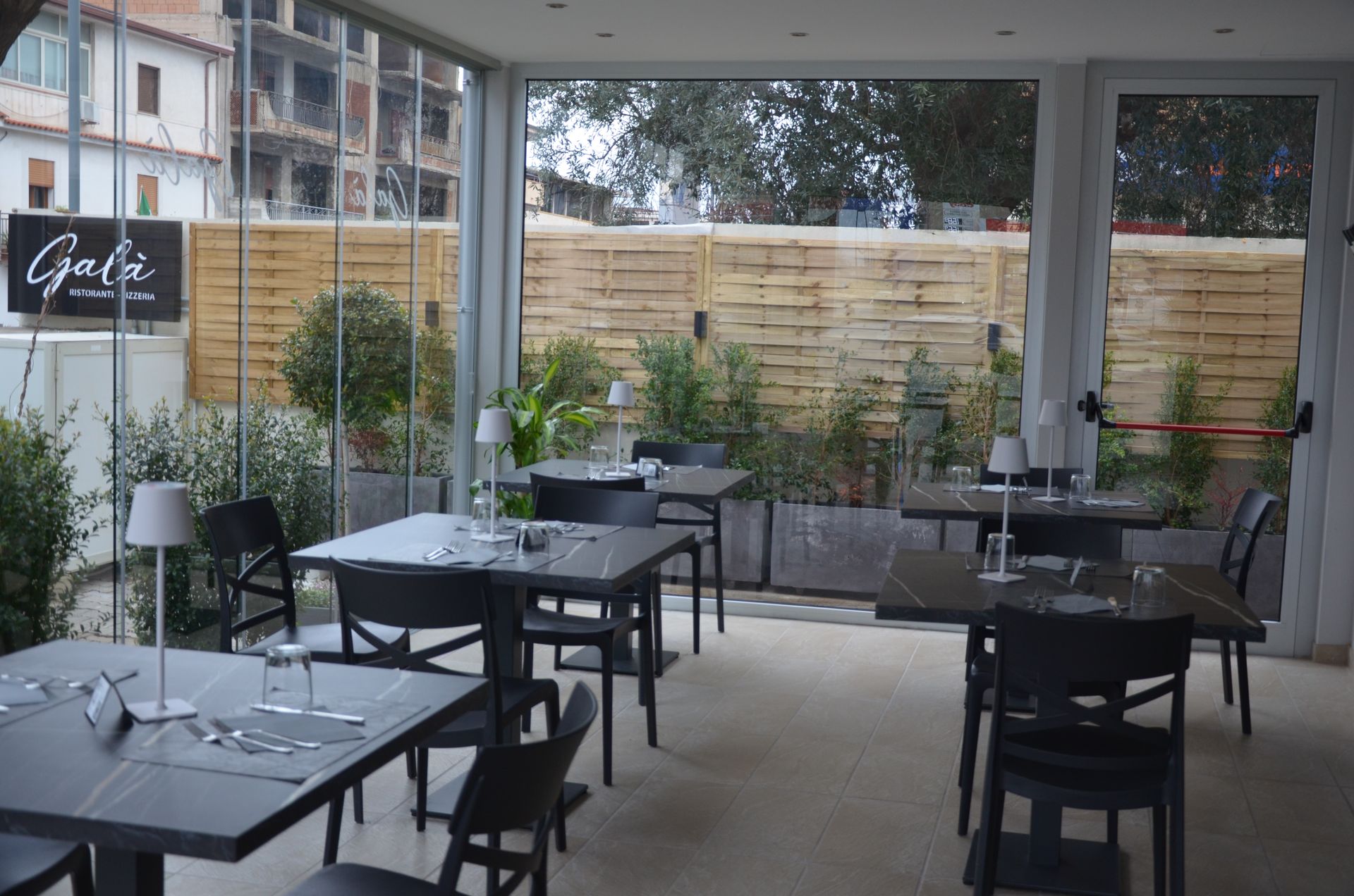 Interno del ristorante con tavoli e sedie vicino alle vetrate, con vista sul verde esterno. Cartello 