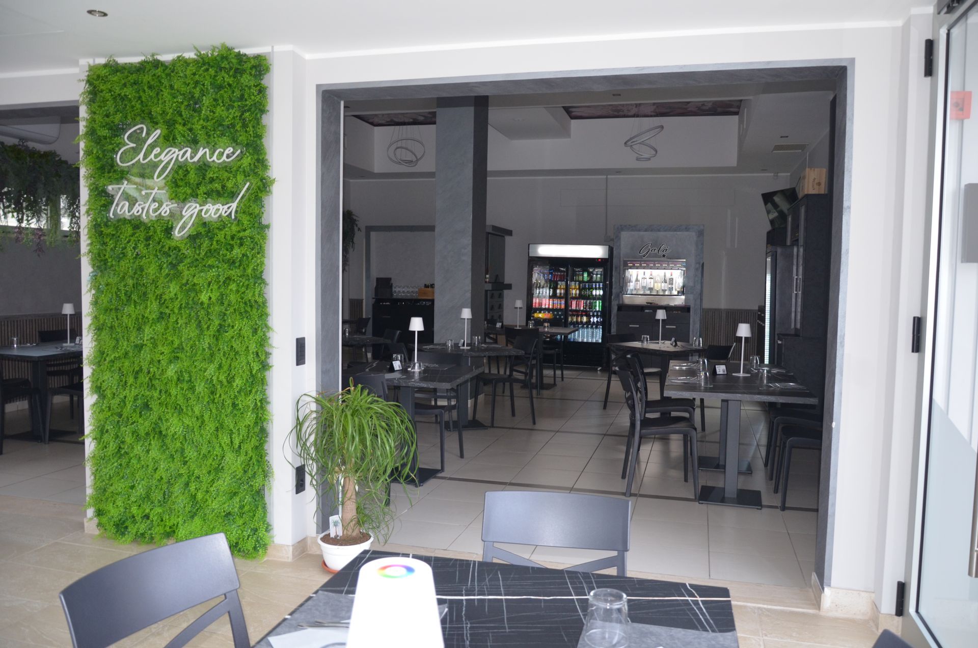 Interno del ristorante con ingresso aperto, parete verde con insegna al neon. Tavoli, sedie e refrigeratore per bevande visibili.