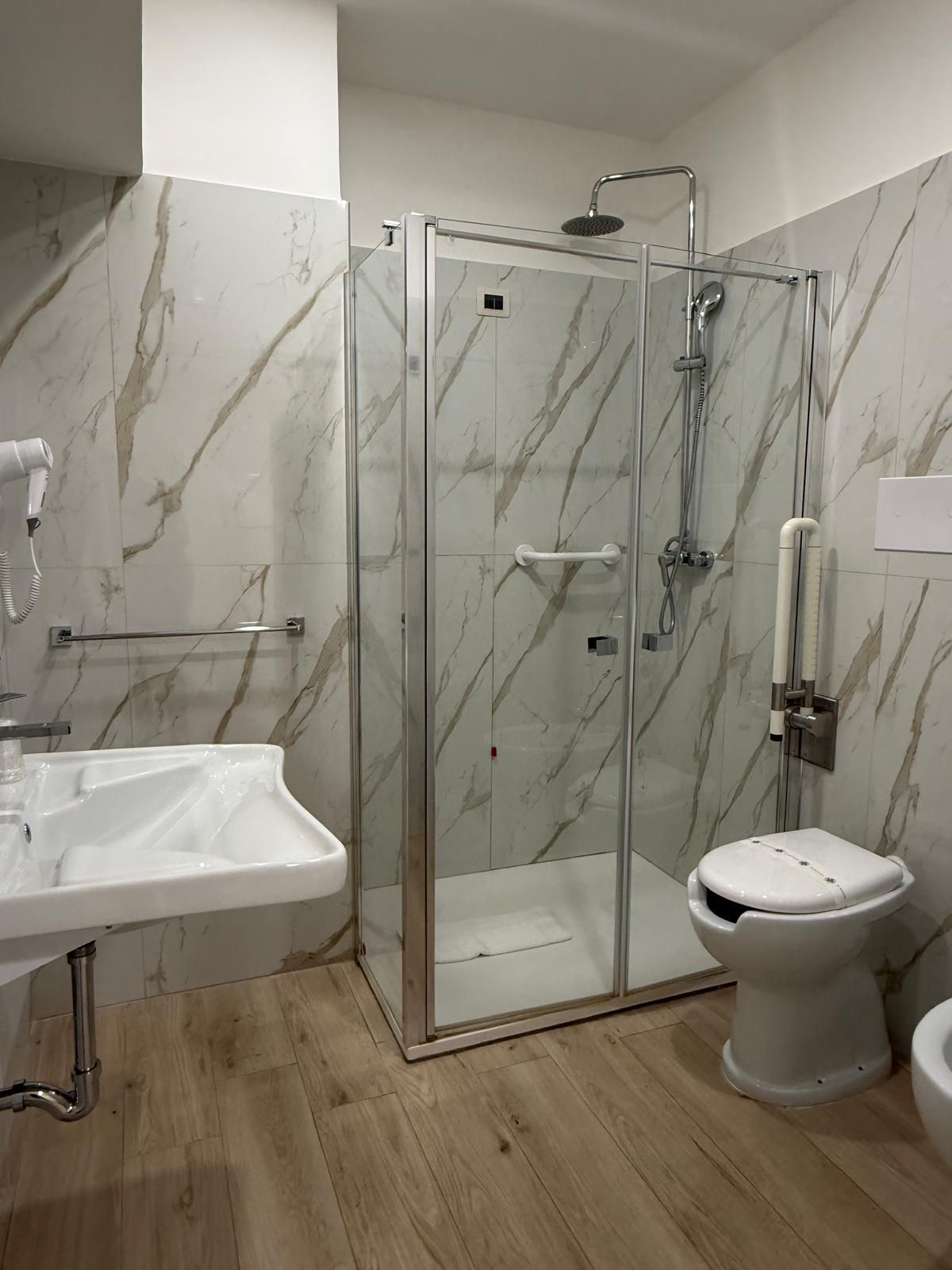 Bagno con lavandino bianco, box doccia, WC e portasciugamani, il tutto con pareti decorate in marmo.