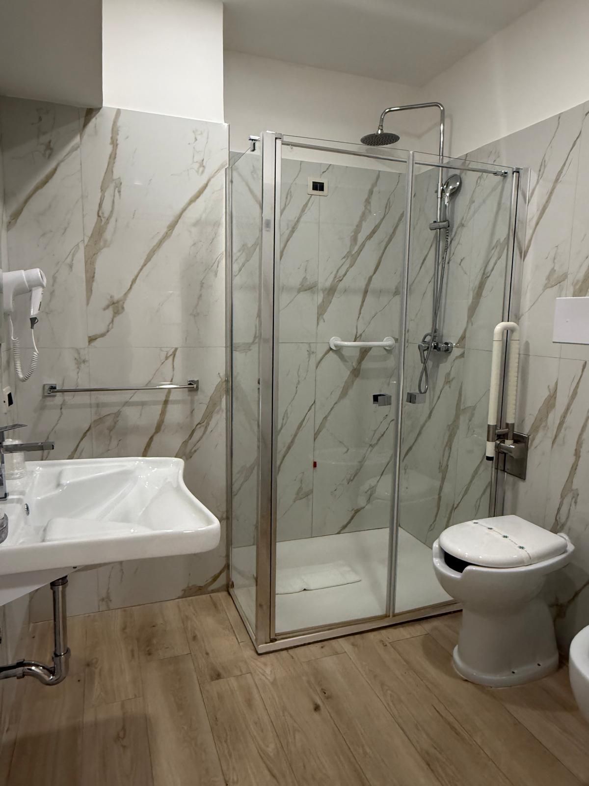 Bagno con doccia in vetro, WC e lavandino, pareti con motivi in ​​marmo e pavimento effetto legno.