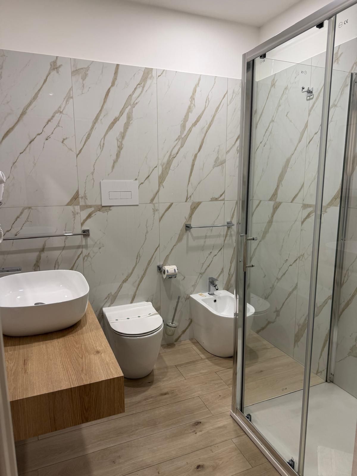 Bagno moderno con piastrelle effetto marmo, mobile lavabo in legno, WC, bidet e porta doccia in vetro.