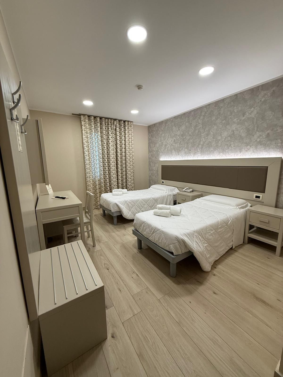 Camera d'albergo con due letti singoli, scrivania e panca, pavimenti in legno chiaro e tonalità neutre.